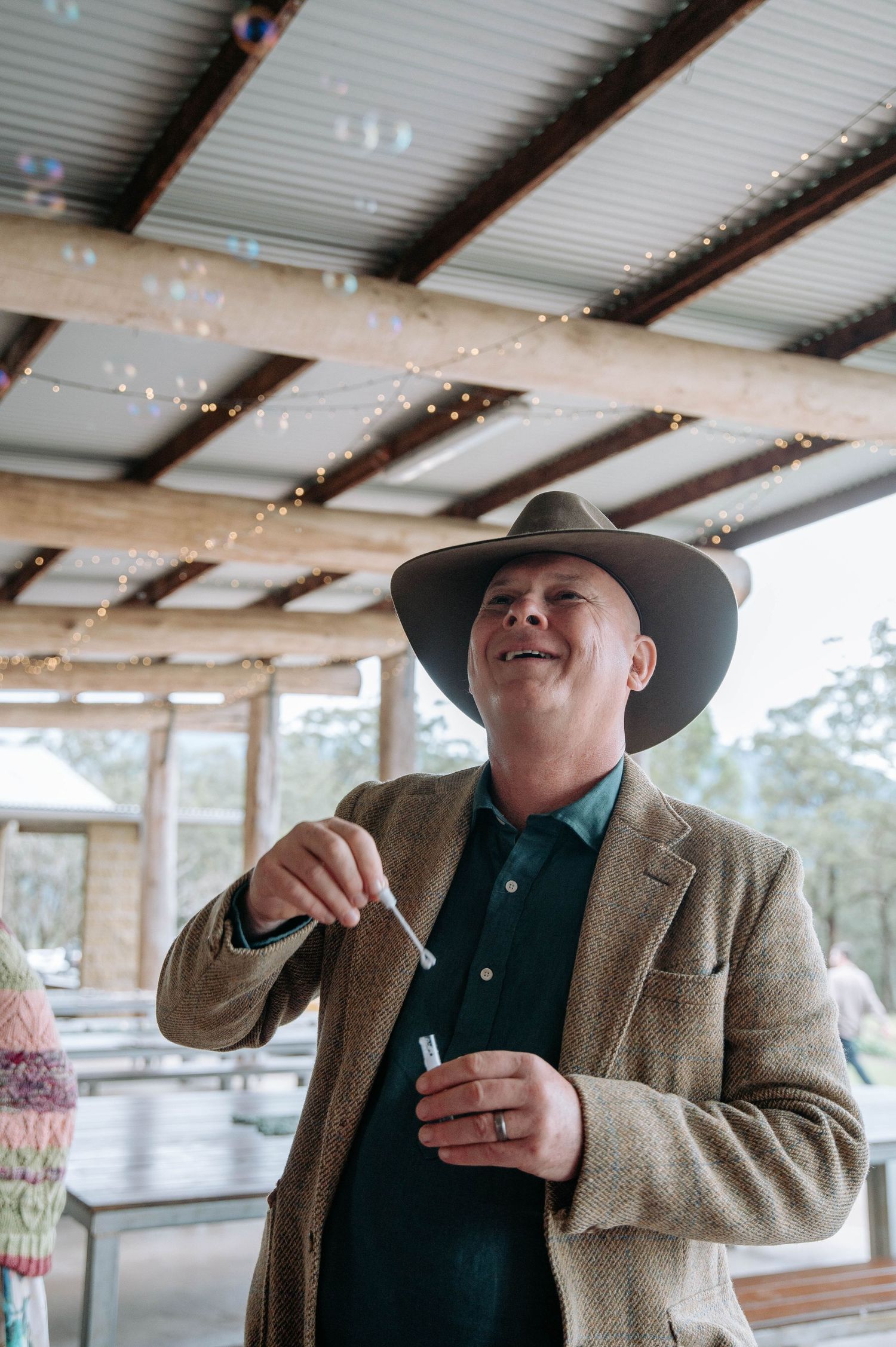 A person in a cowboy hat and blazer laughs joyfully while standing under a metal roof structure.