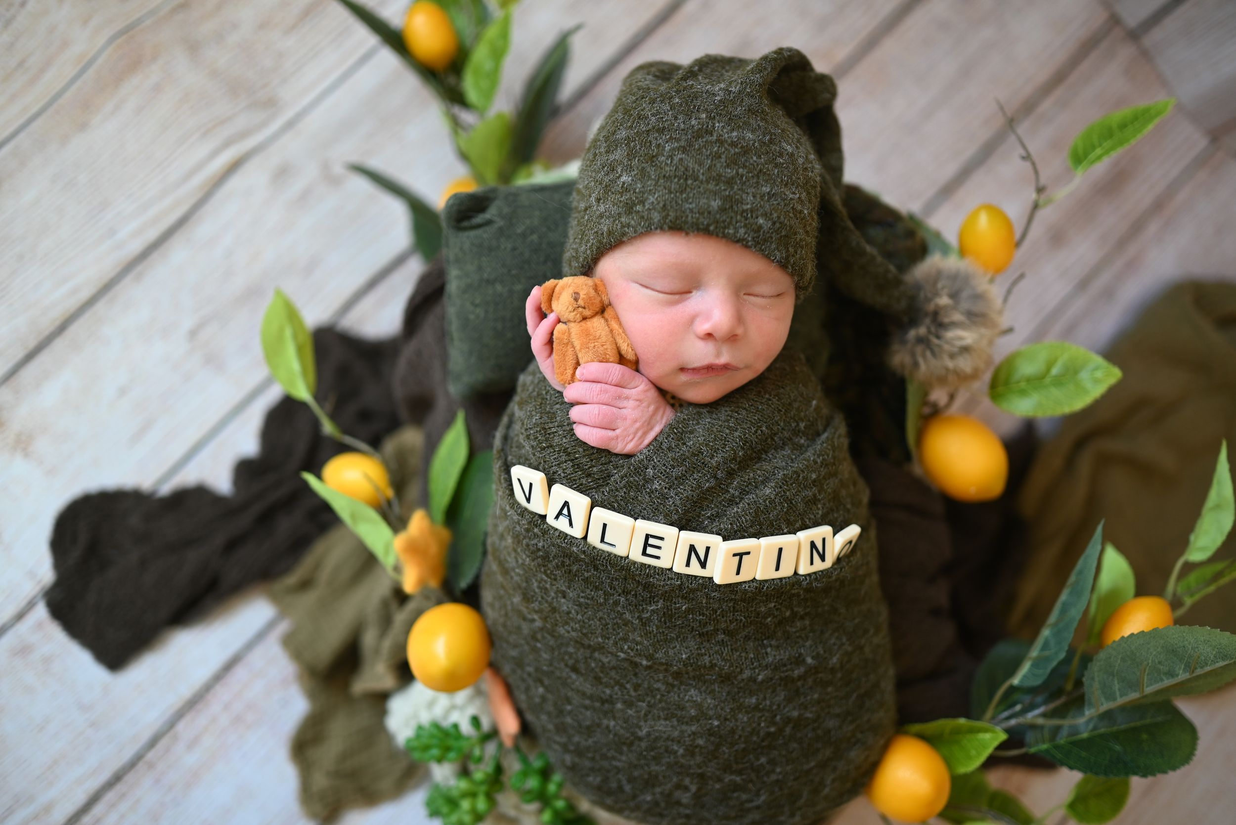 Newborn swaddled in dark green with yellow citrus decor styled on whitewashed wood background.