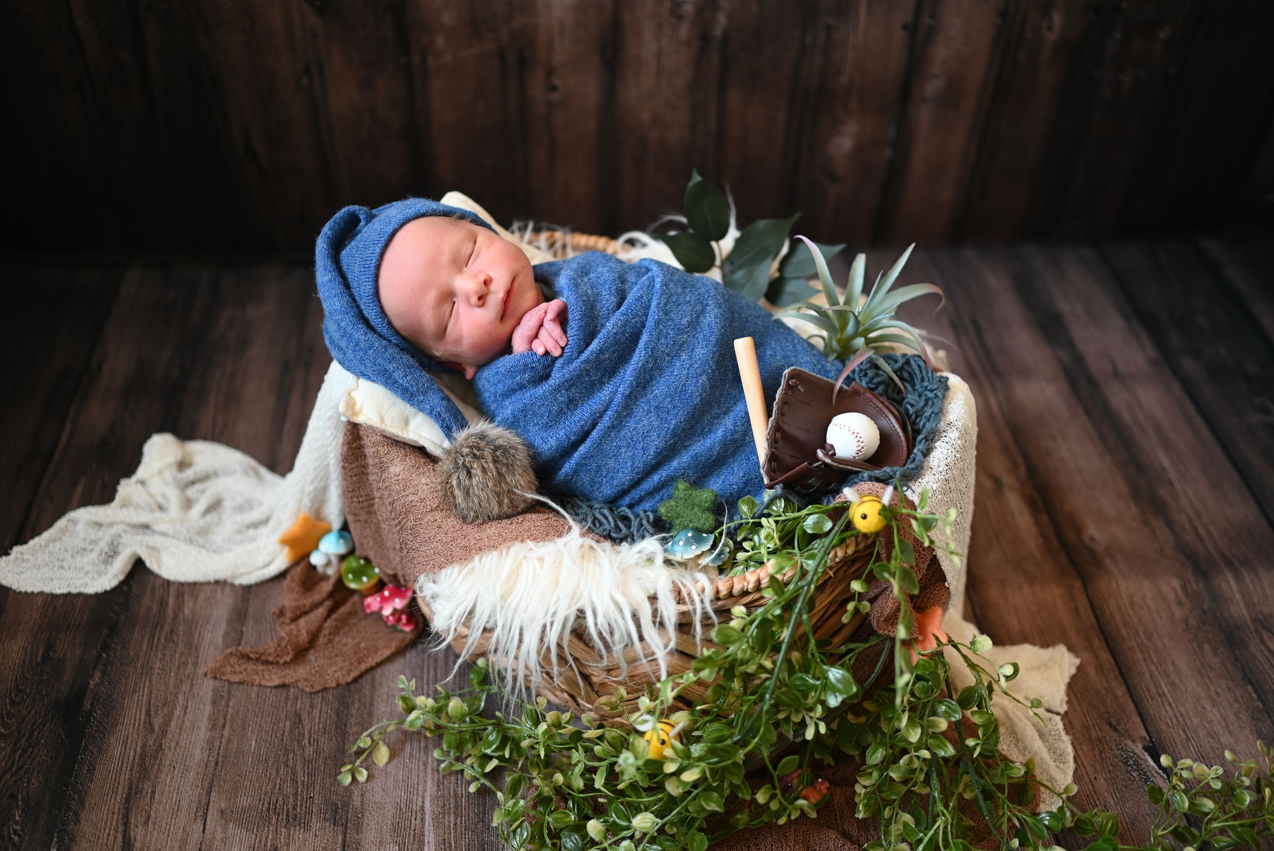 A sleeping newborn baby wrapped in a blue blanket rests in a rustic basket with greenery and flowers on wooden flooring.