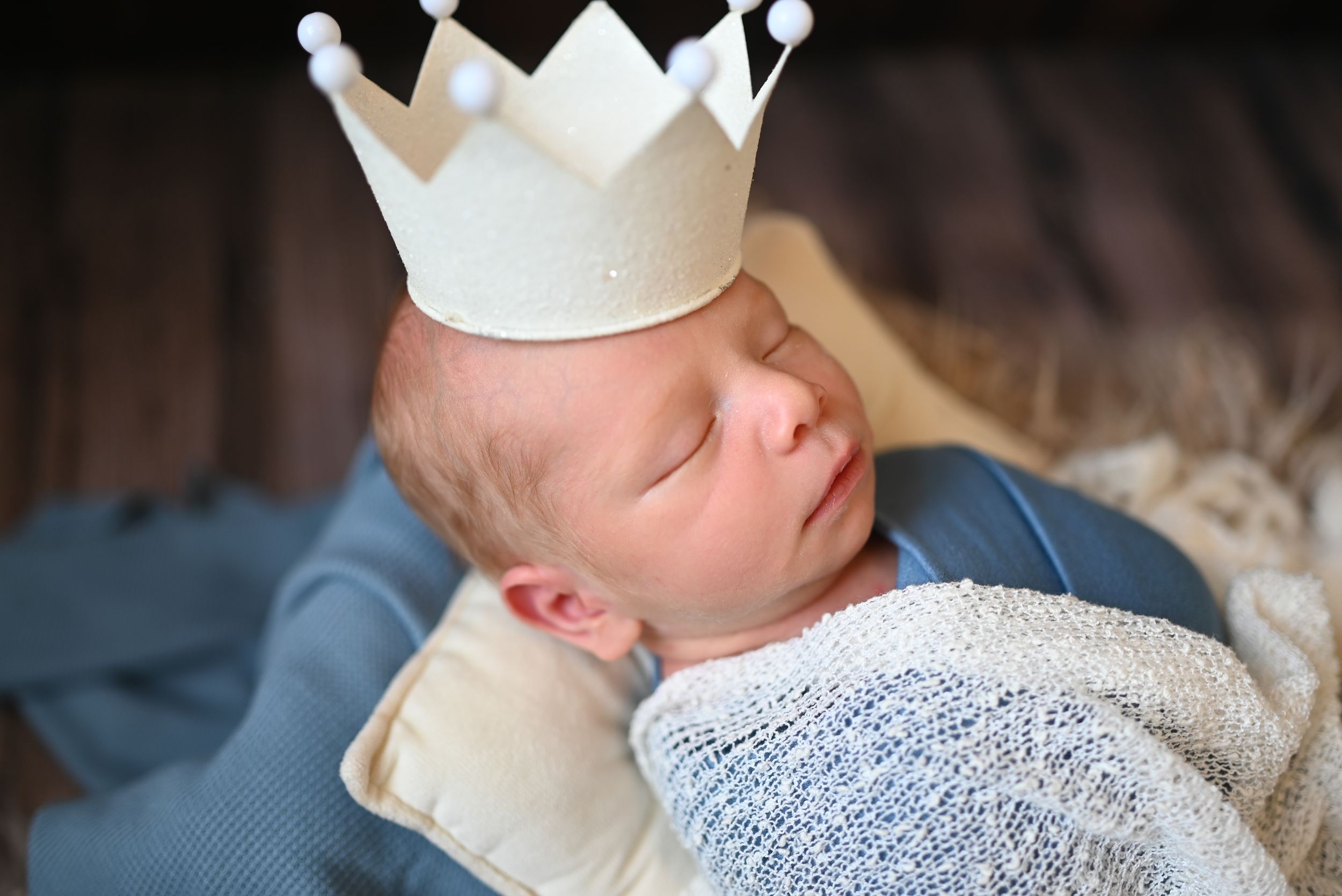 A baby in a crown sits among cozy blankets and pillows, creating a regal portrait setting.