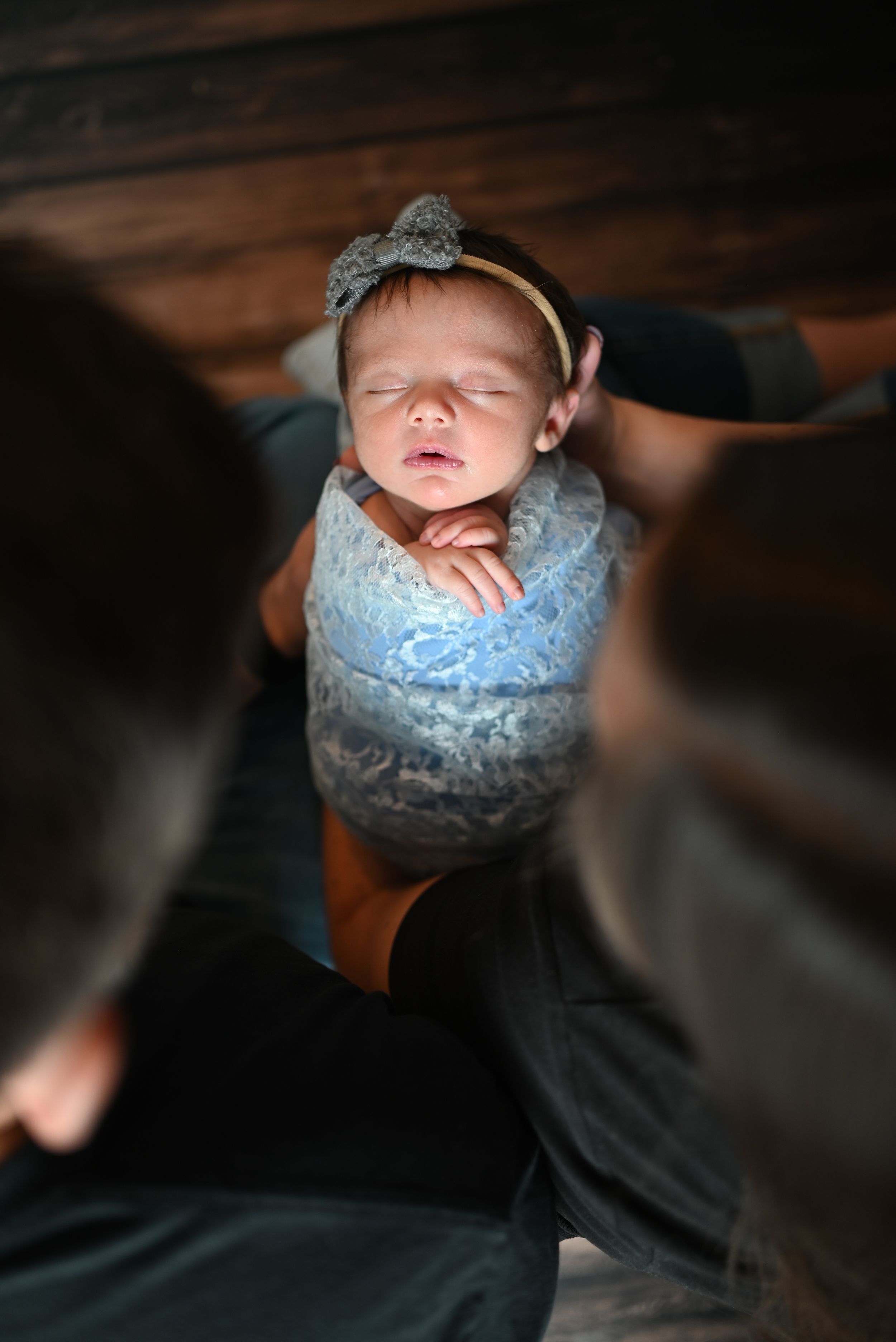 Sleeping newborn wearing soft blue lace outfit and gray bow headband in moody portrait lighting.