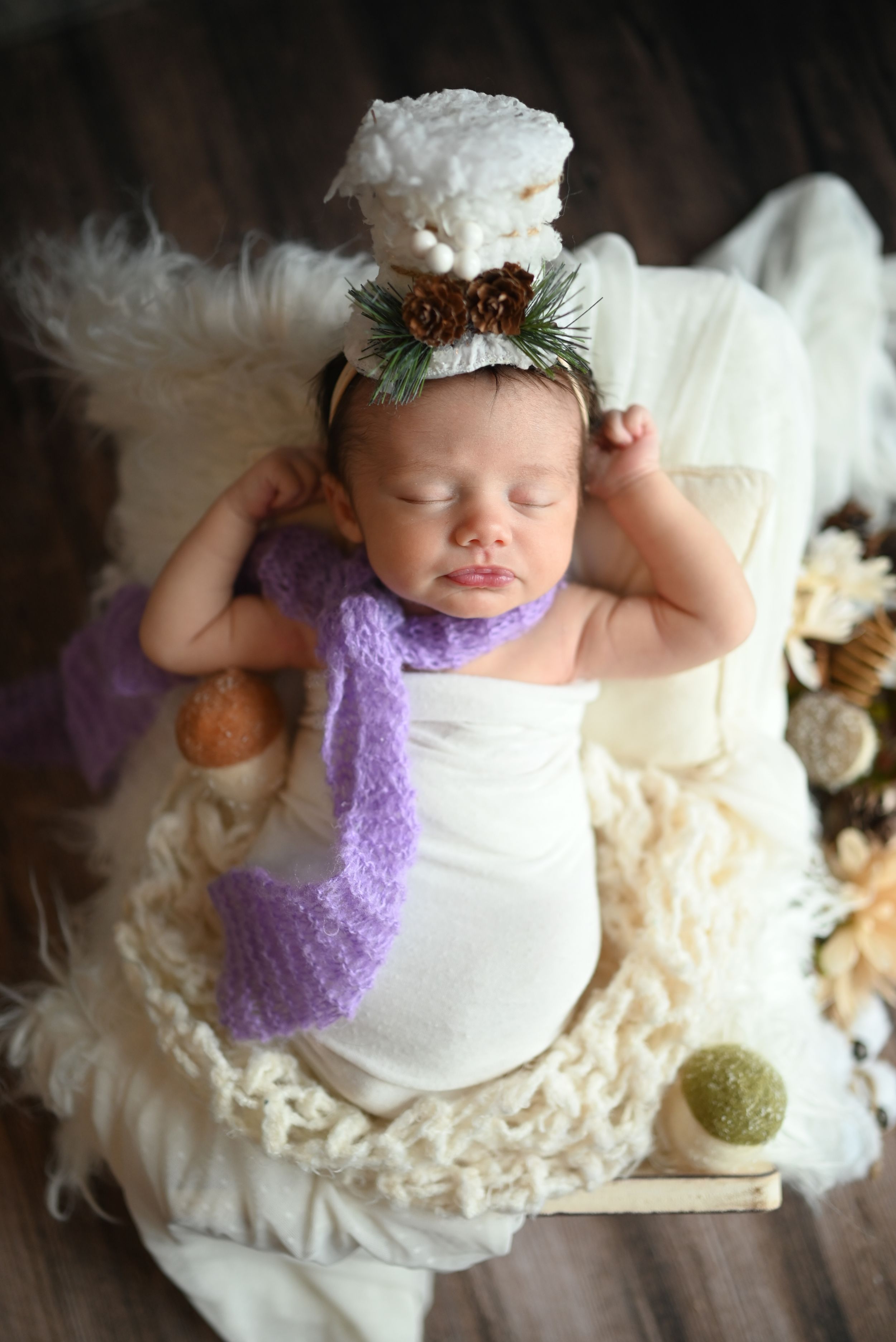 A sleeping baby wearing white clothing and lavender accessories rests on a cream-colored textured blanket.