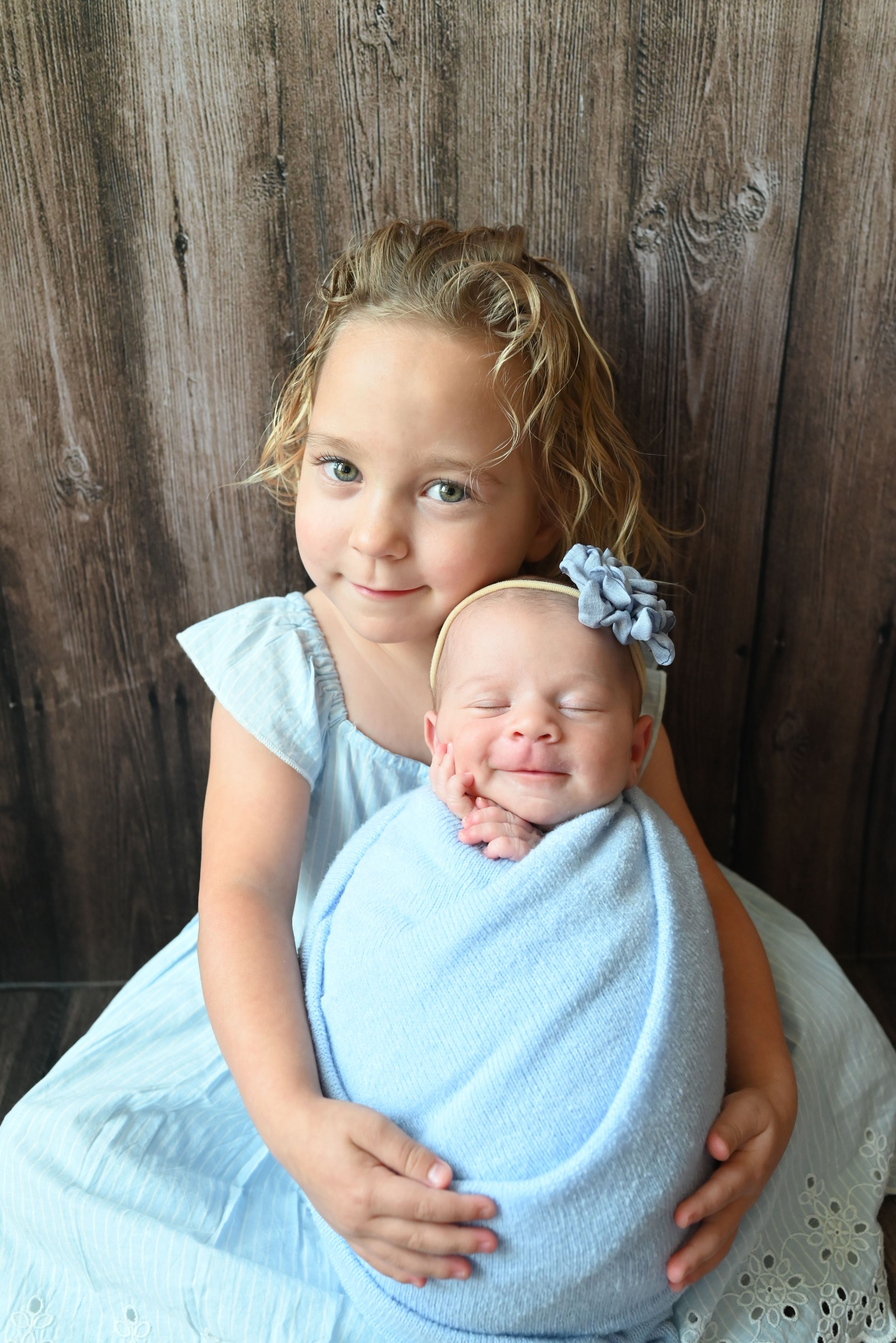 Two children with light hair sitting together against a rustic wooden fence background.