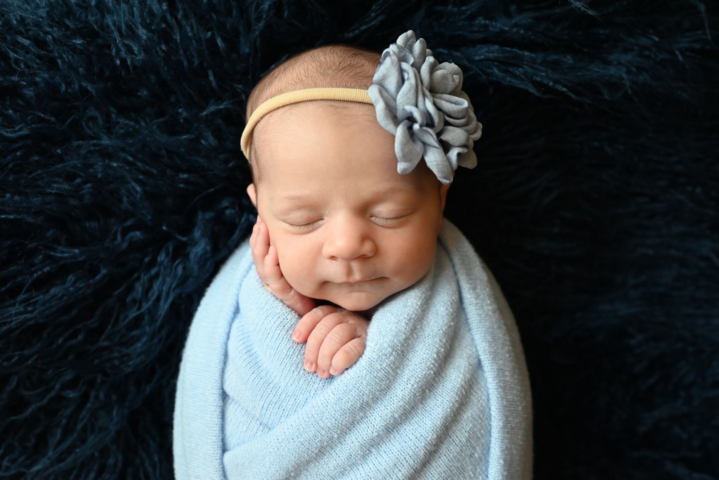 A sleeping newborn swaddled in light blue blanket with silver flower headband on dark fur backdrop.
