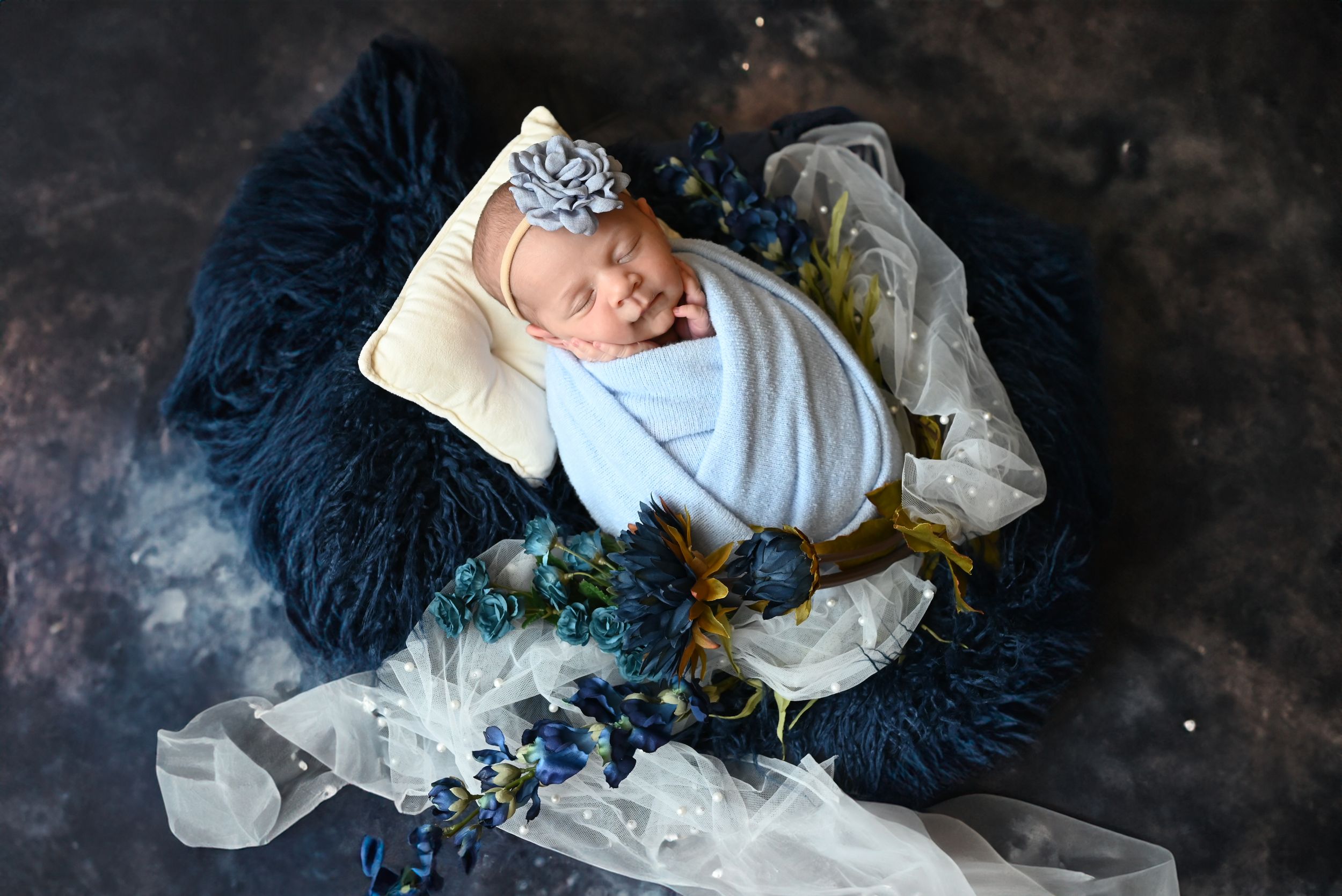 Sleeping newborn baby wrapped in white swaddle with crown, resting on navy blue and white textiles.