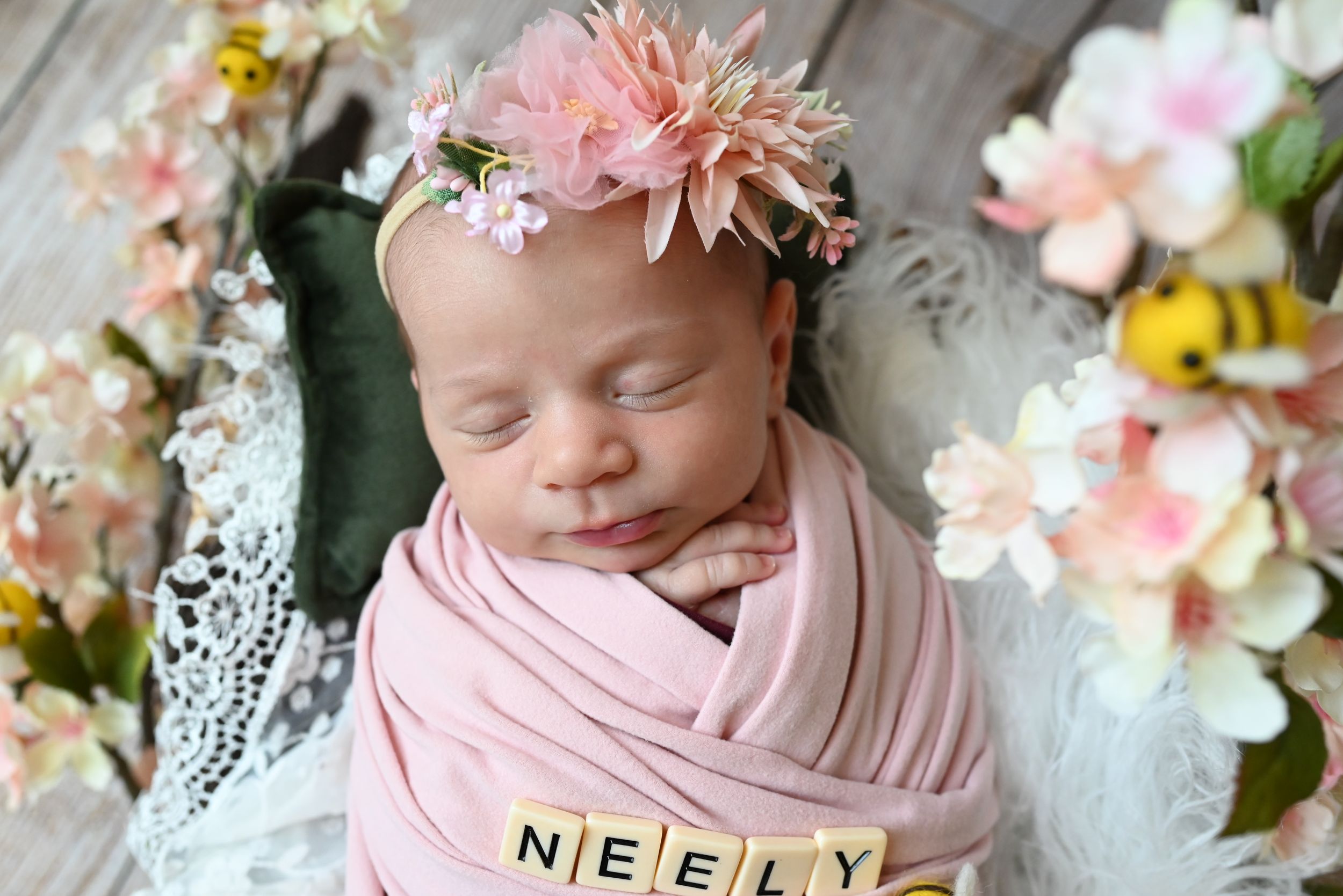 A sleeping baby wrapped in pink swaddle with floral crown amid spring blossoms and wooden letter blocks.
