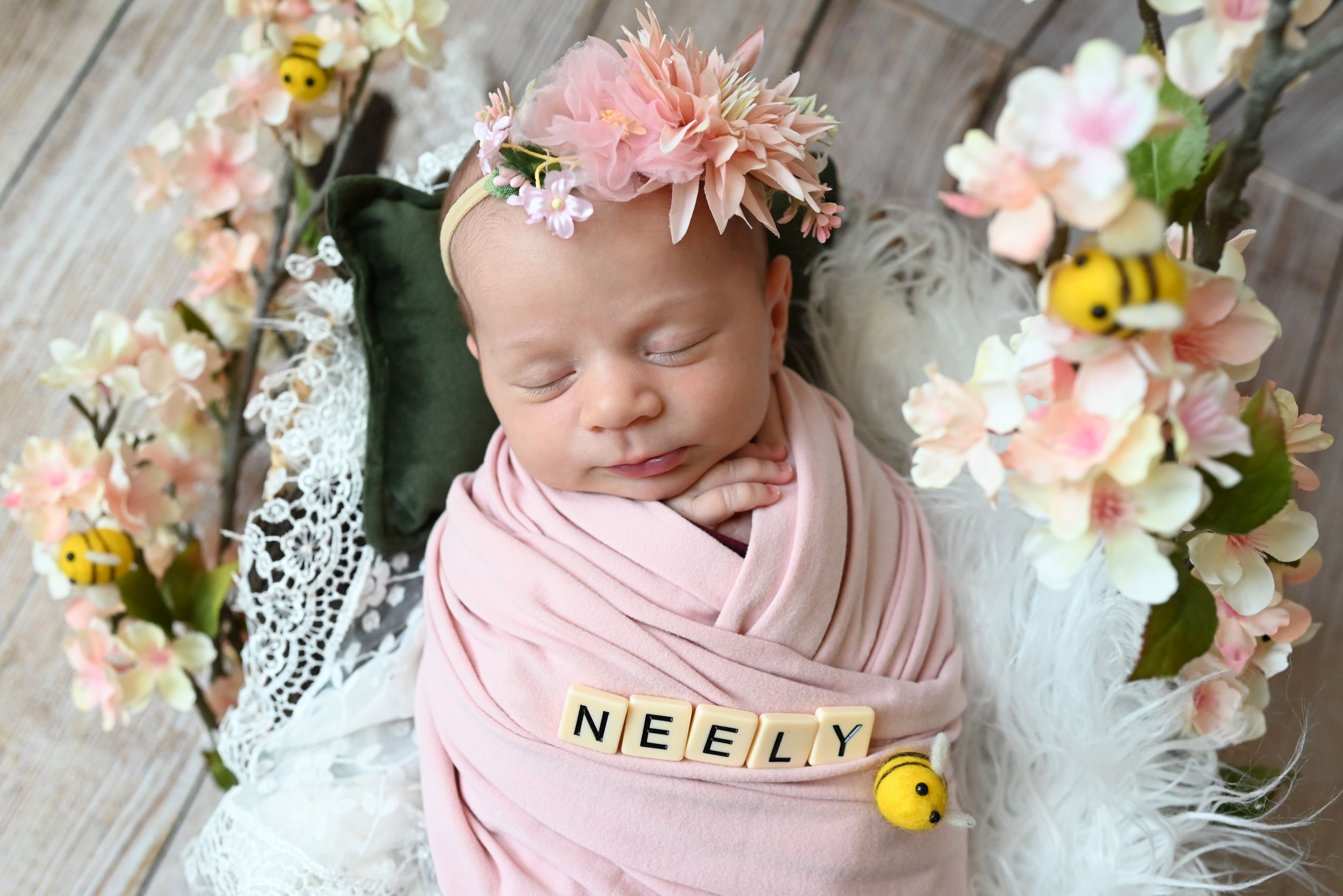 Newborn baby swaddled in pink fabric with letter blocks spelling NEELY surrounded by pink and white flowers on lace backdrop.