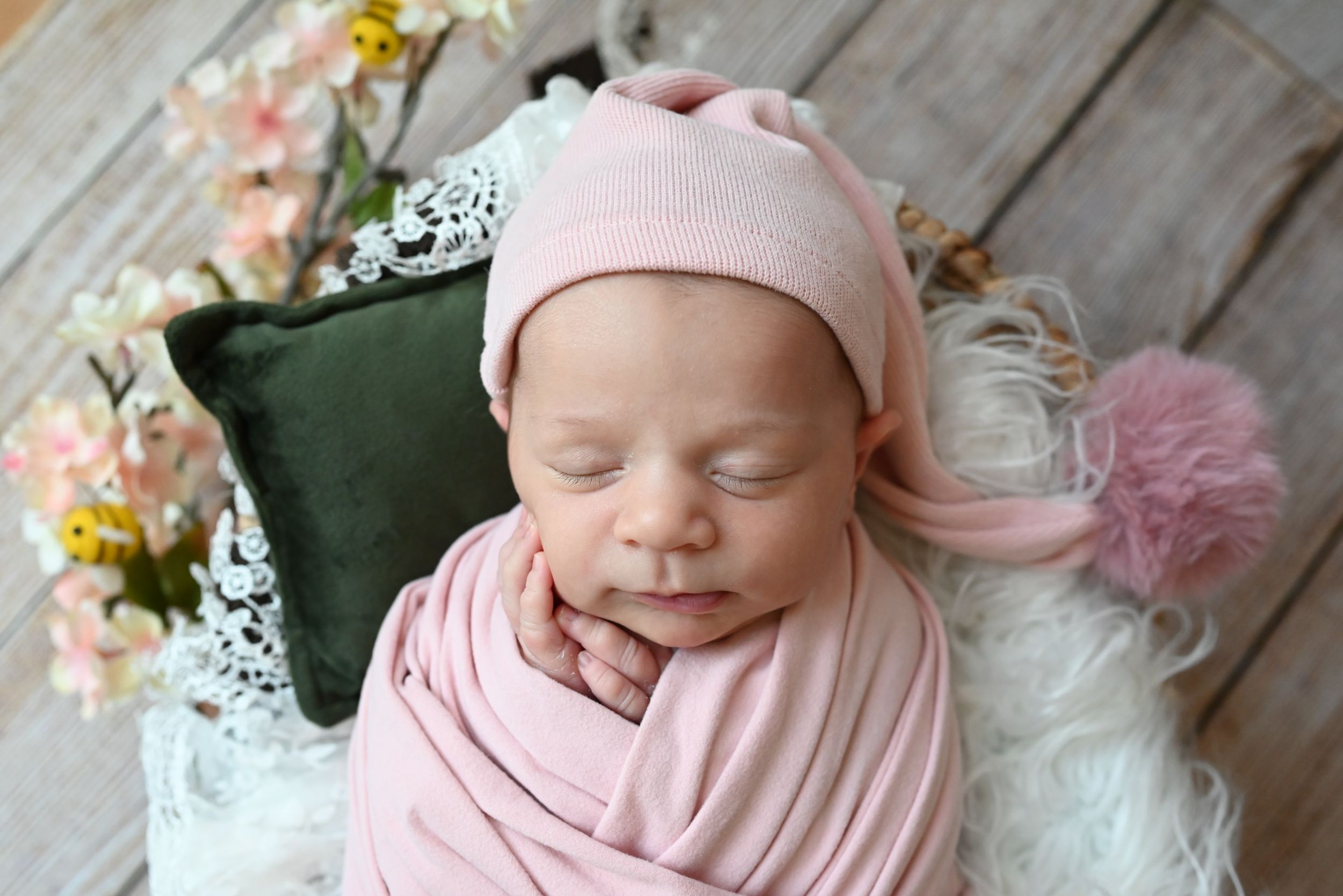 A newborn baby swaddled in soft pink fabric sleeping peacefully on white lace with floral decorations.