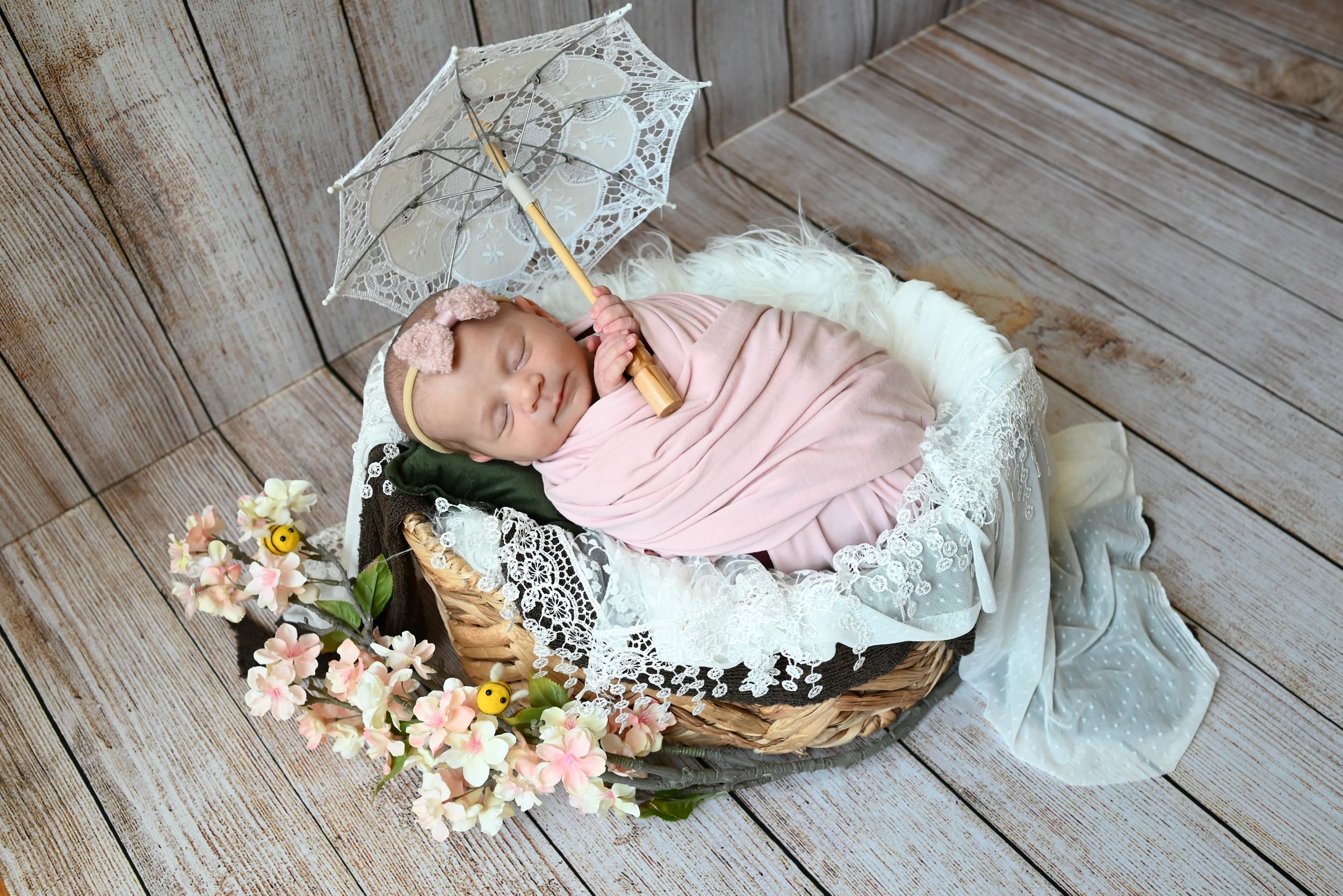 Newborn sleeps peacefully in wooden basket with lace umbrella, pink wrap, and floral decorations on wooden floor.