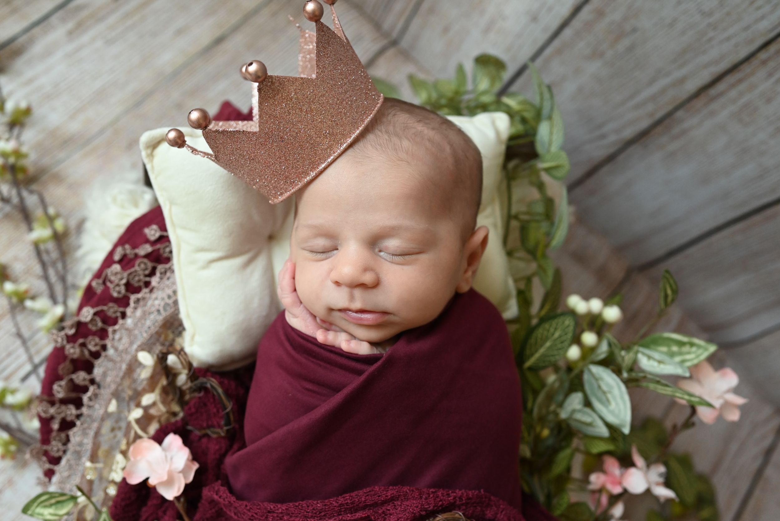 Adorable newborn baby sleeping in a wooden bed surrounded by soft pink and white flowers and greenery.