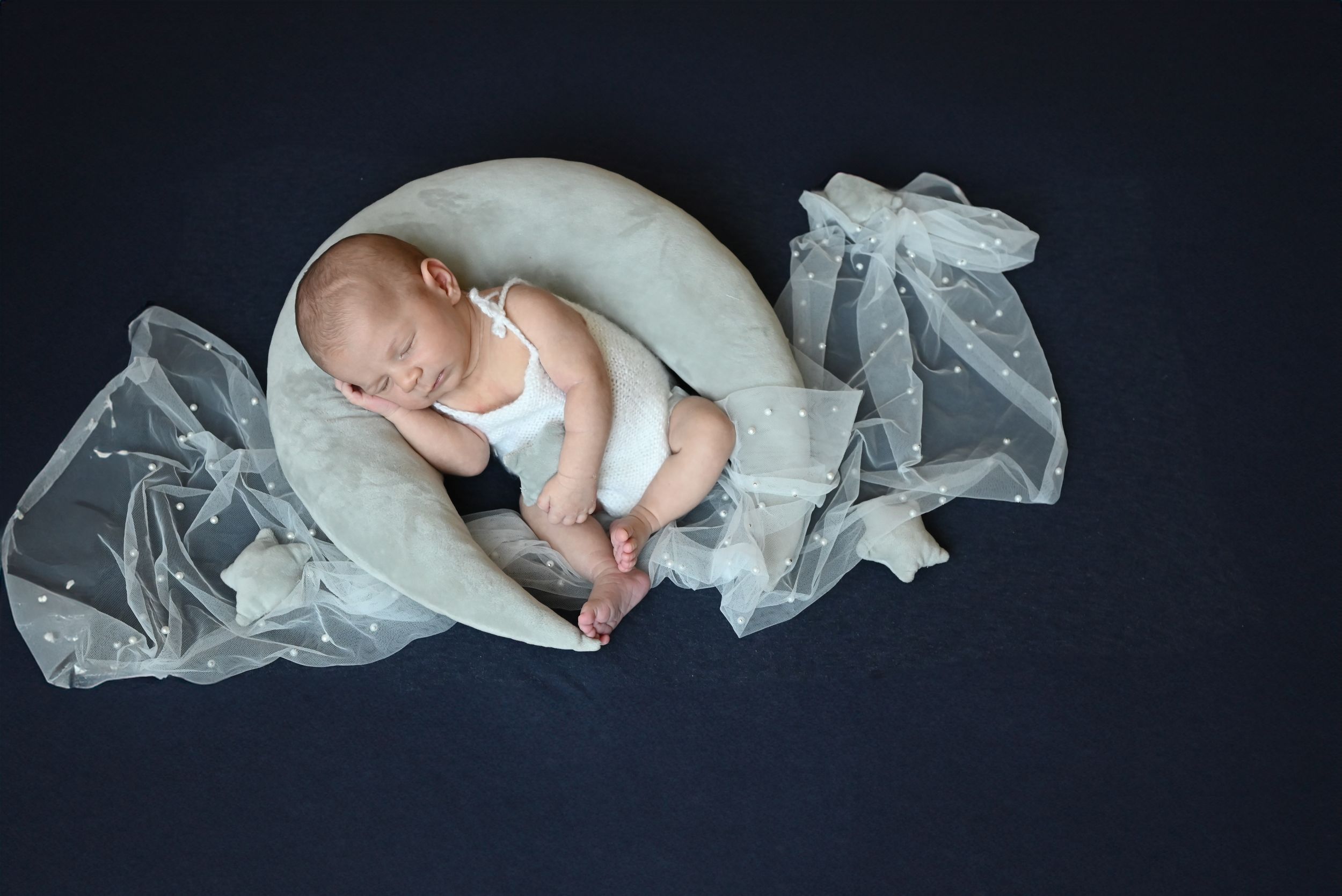 Newborn baby sleeping peacefully on a crescent moon prop with flowing tulle fabric on dark background.