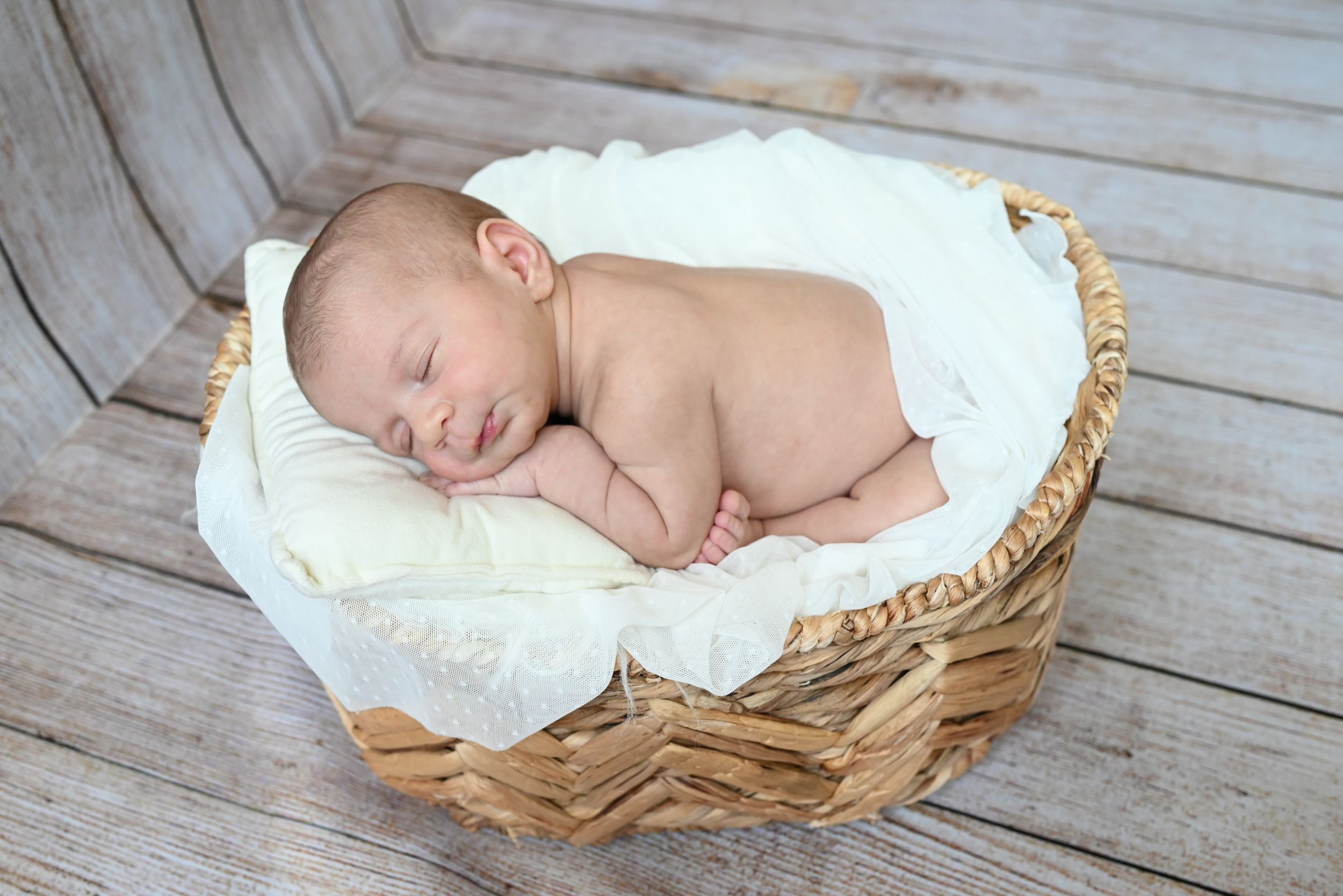A sleeping newborn rests peacefully on soft white fabric in a rustic woven basket on wooden floor.