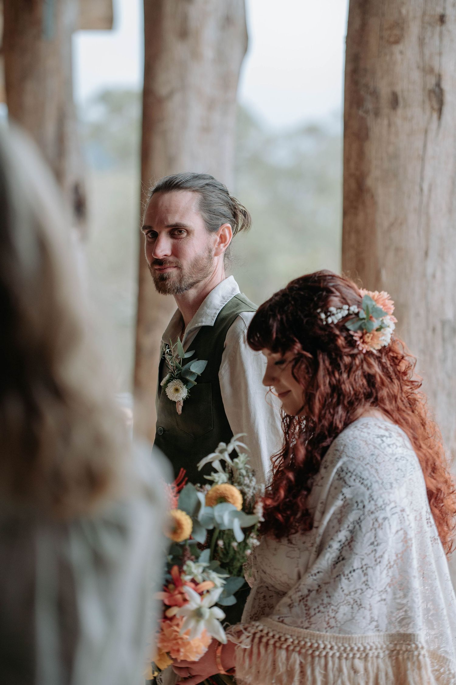 A romantic outdoor wedding ceremony featuring a lace-clad bride and groom standing among rustic wooden pillars.
