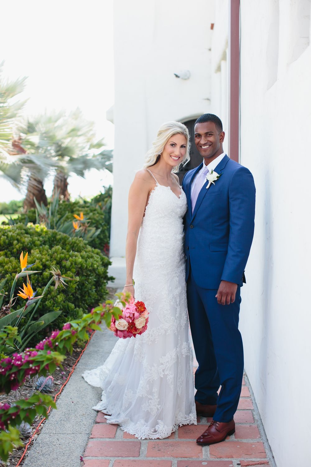 A couple in wedding attire poses on a white columned steps surrounded by tropical flowers and greenery.