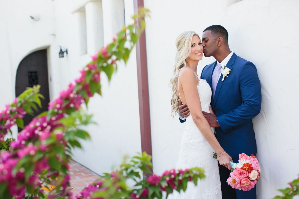 Newlywed couple shares intimate moments against a white wall with bright pink bougainvillea flowers.