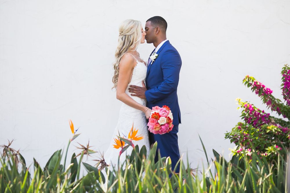 A couple in wedding attire embraces in a tropical garden with pink bougainvillea flowers and birds of paradise plants.