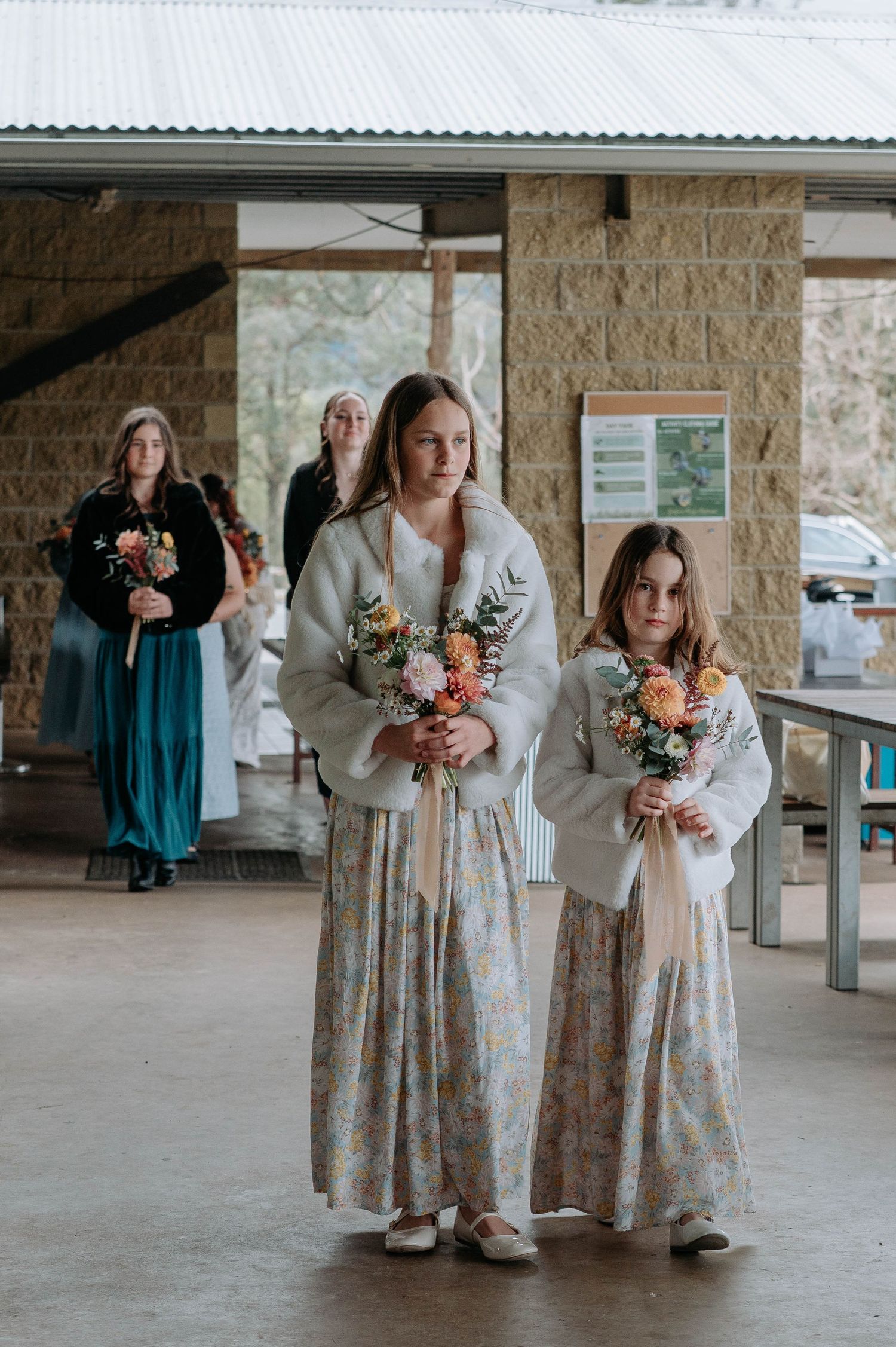 Bridesmaids in floral dresses and cozy cardigans hold autumn-colored bouquets while posing under a covered walkway.