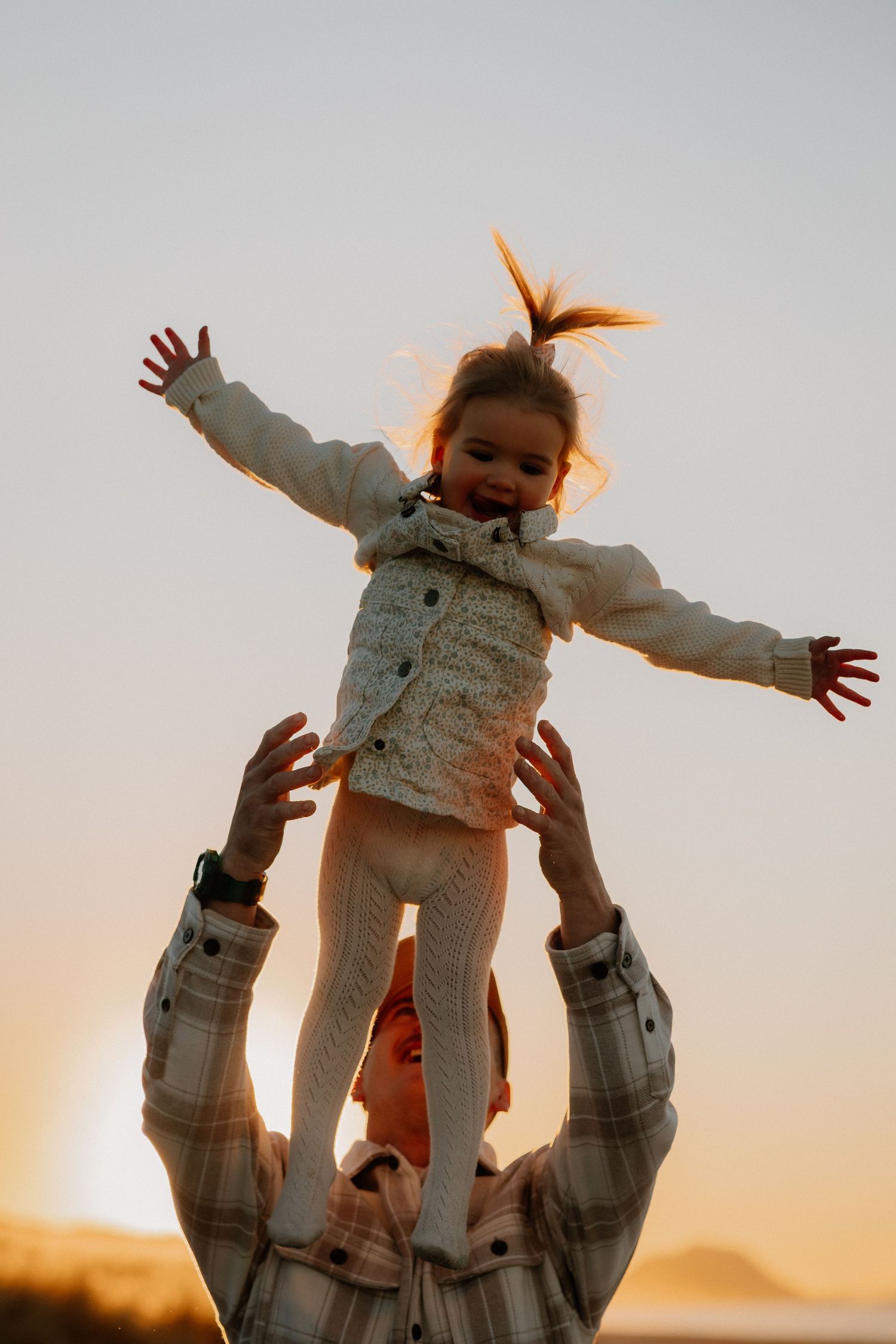 A joyful child is tossed into the air against a sunset sky, wearing white clothing and spreading arms wide.