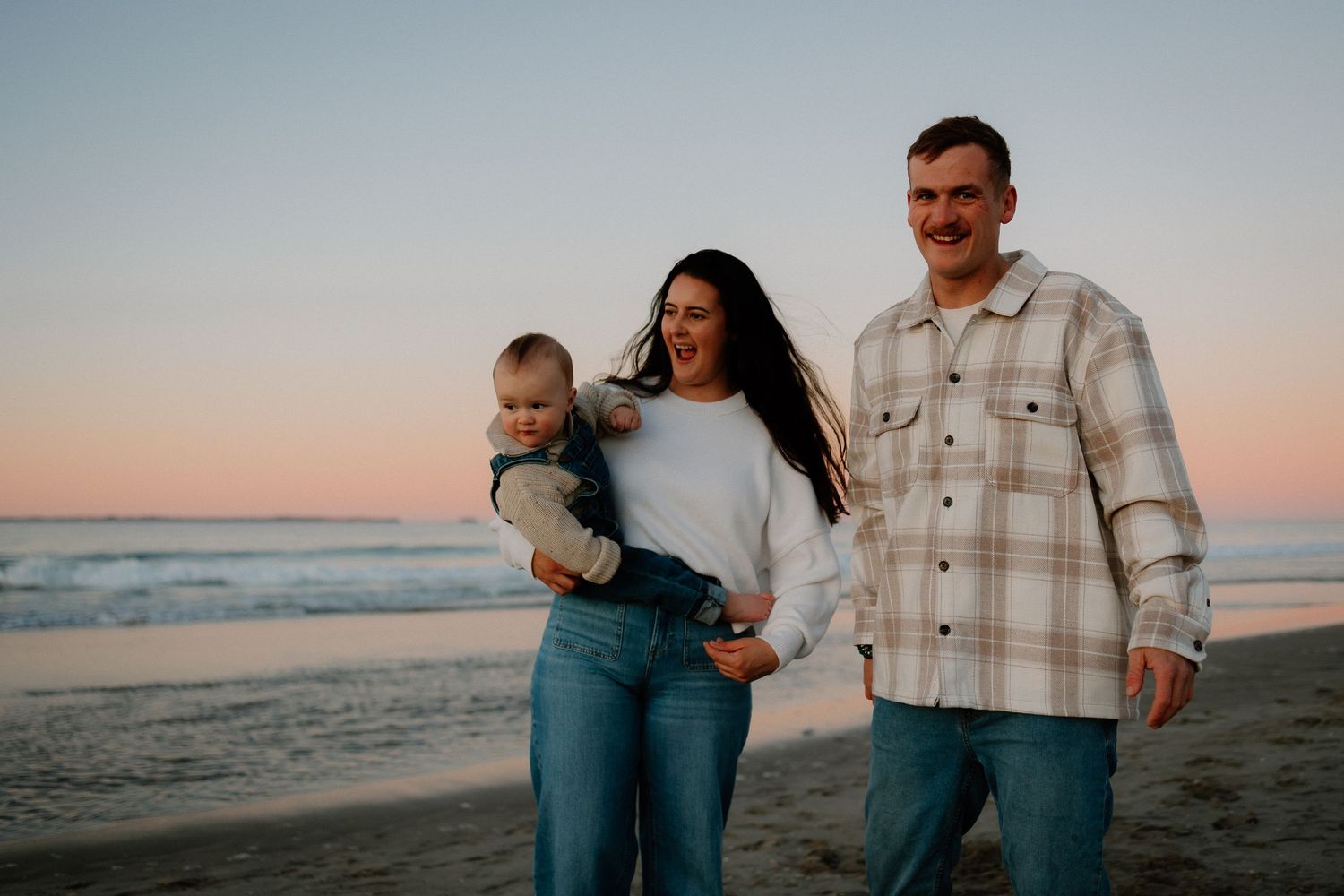 A family walks together on the beach at sunset while wearing casual attire against the ocean backdrop.
