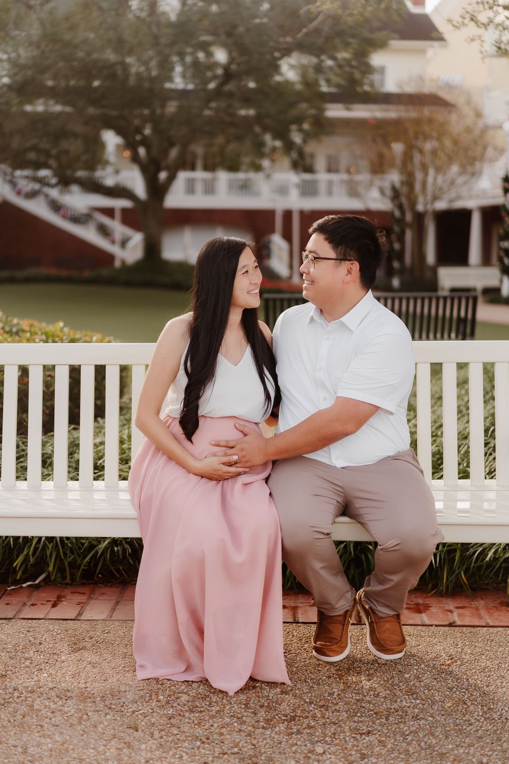 A couple in white and pink outfits share romantic moments on a park bench during an outdoor photoshoot.
