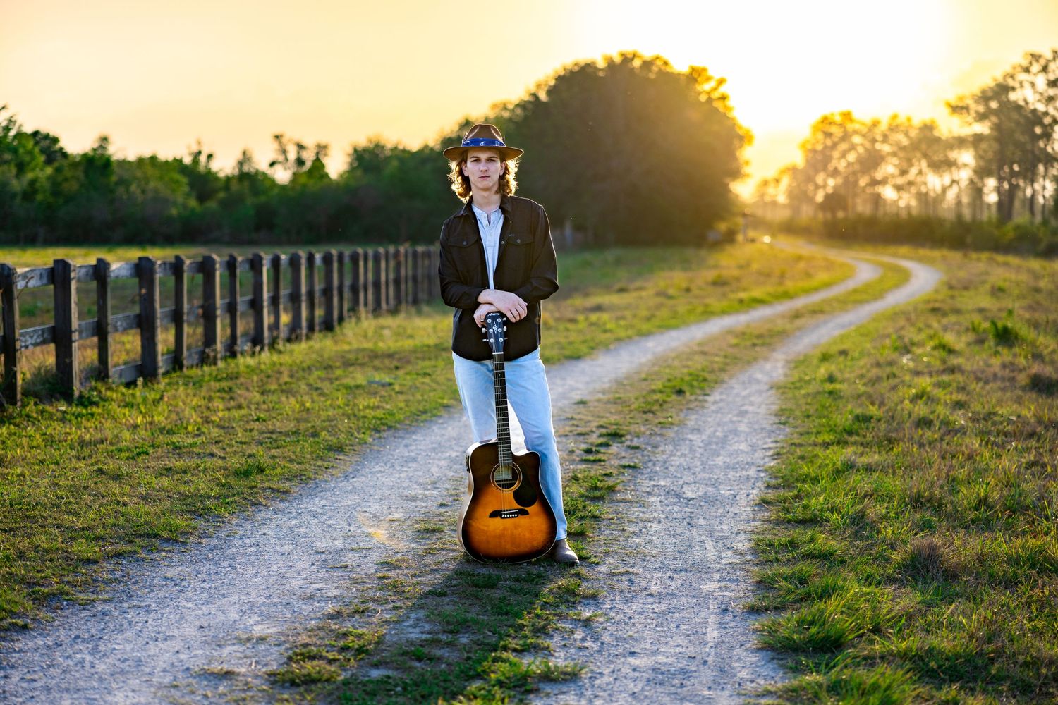 A scenic country pathway at golden hour with tall wooden fence posts leads into warm sunset light.