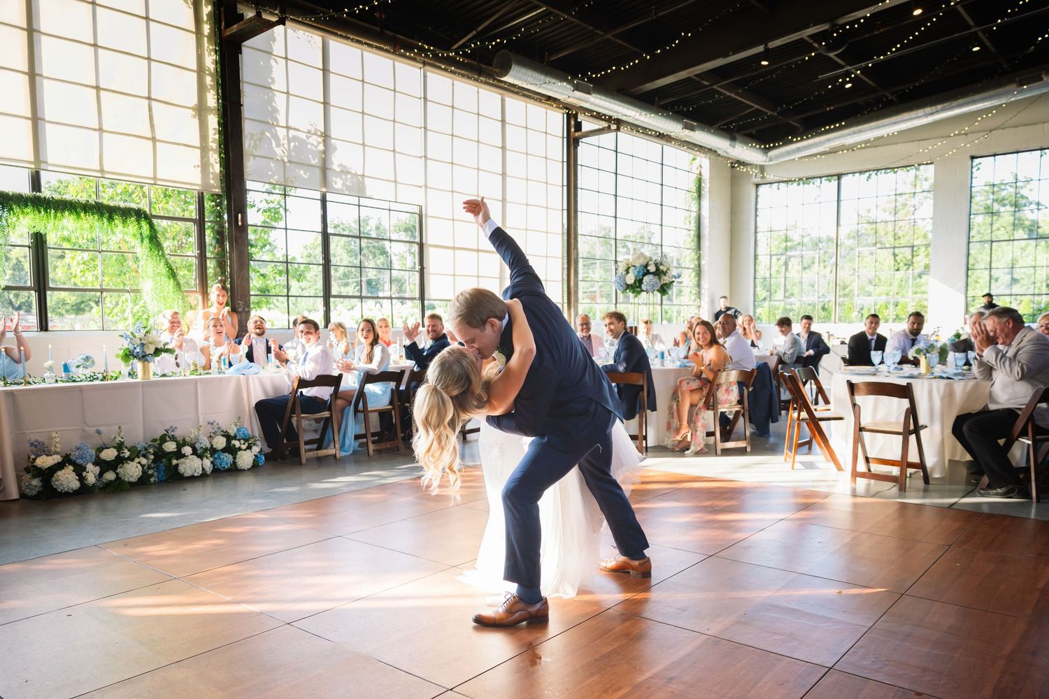 A couple performs a romantic dip while dancing together at a light-filled reception venue with guests watching in background.