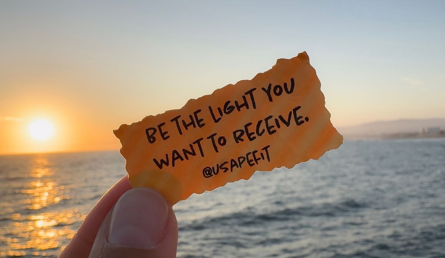 Inspirational quote on torn paper held against a scenic ocean sunset view with golden sunlight reflecting on water.