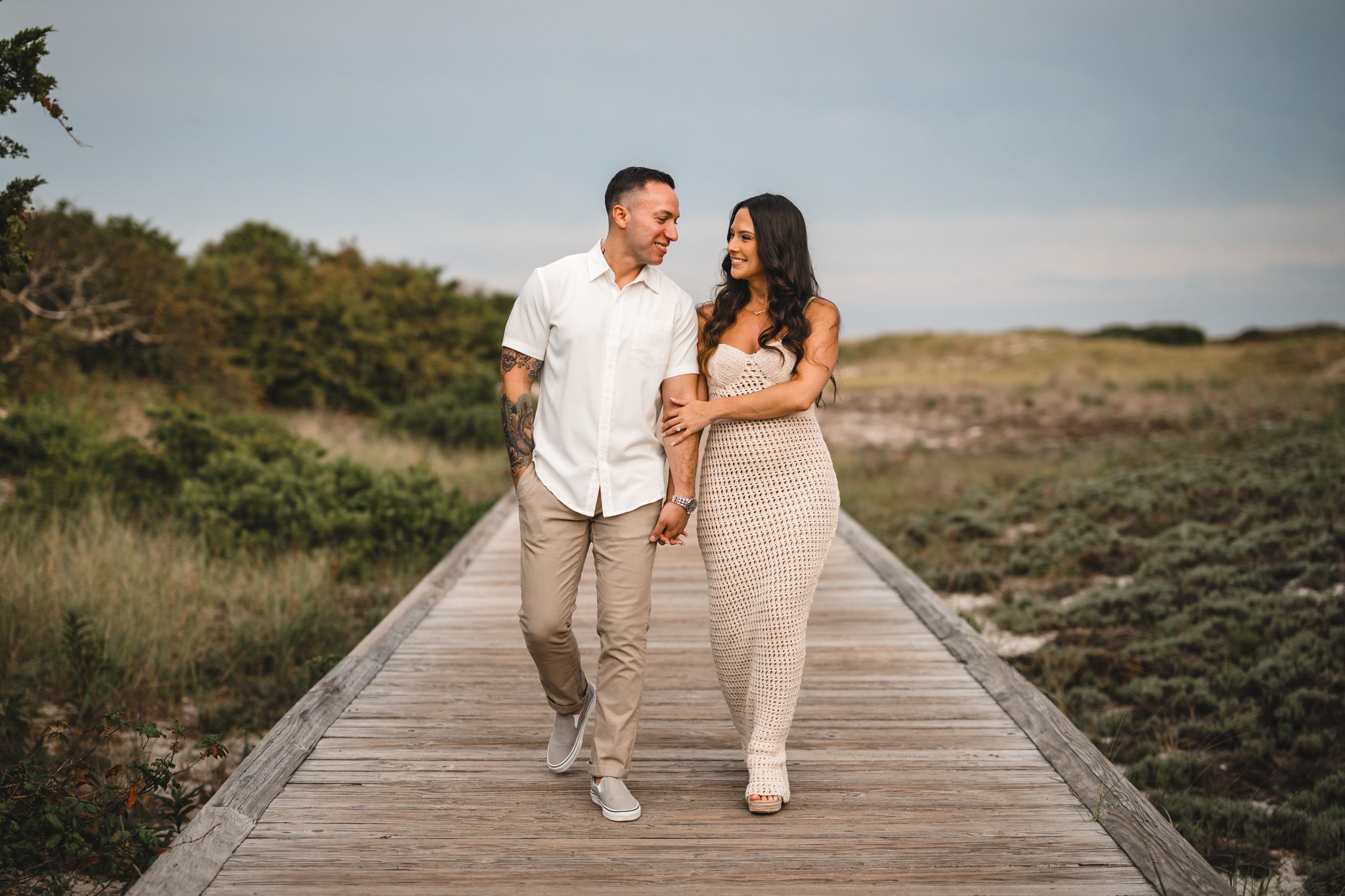 A couple walks along a wooden boardwalk by the beach during golden hour.
