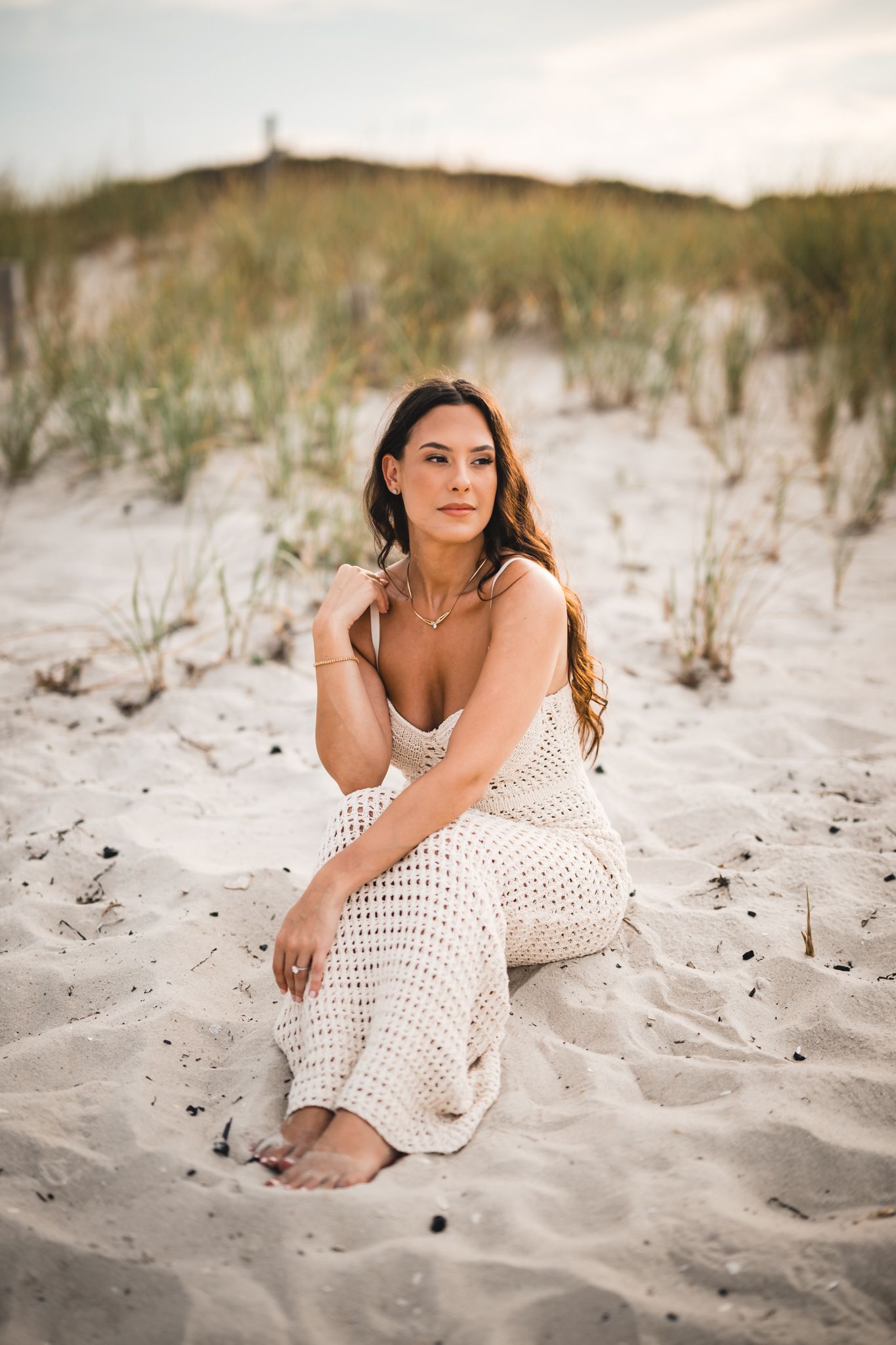 Woman in white crochet dress poses gracefully on sandy beach during sunset.