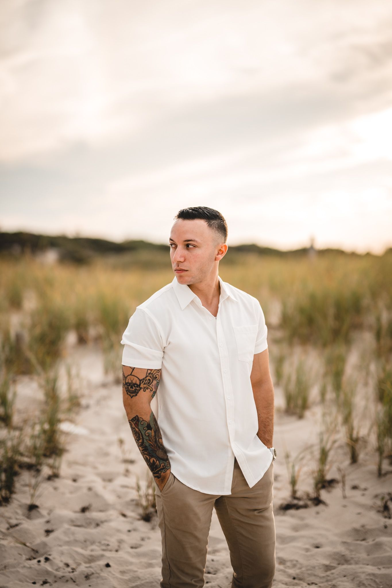 Person in white shirt and khaki pants stands on sandy beach with grass in background.