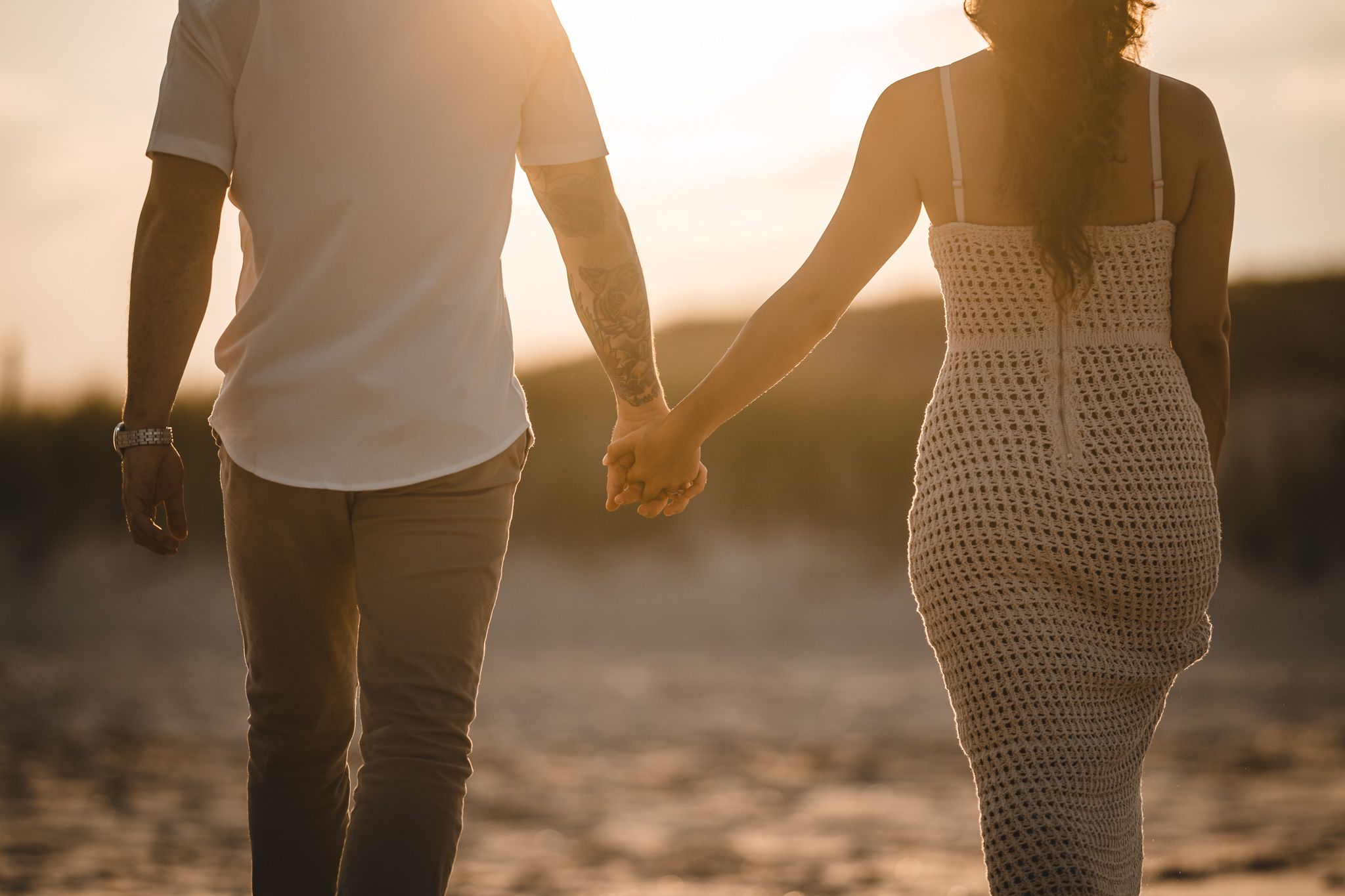 Couple walking hand in hand on sandy beach during golden sunset.