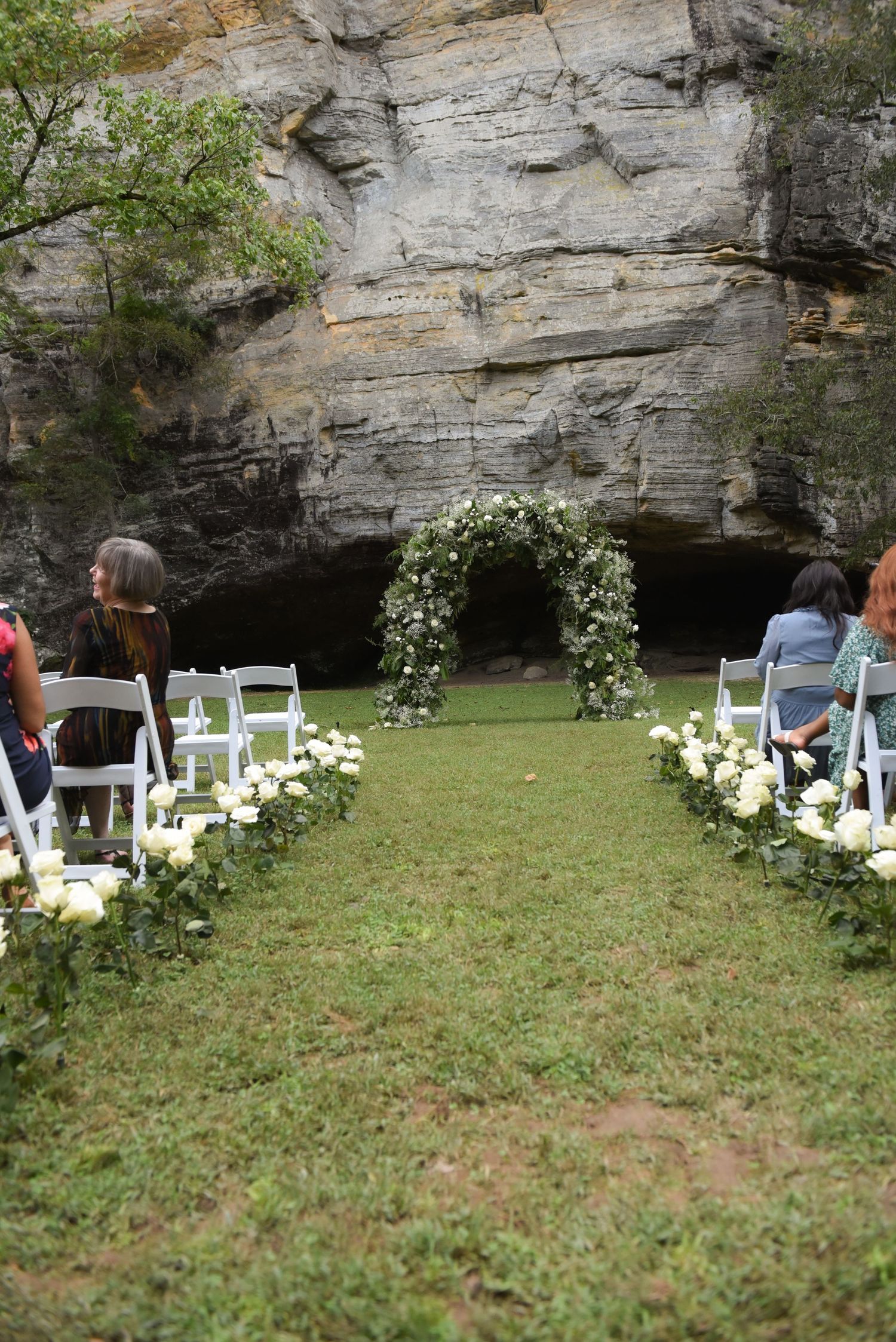 White chairs and floral arrangements set up for an outdoor wedding ceremony against a rock wall backdrop.