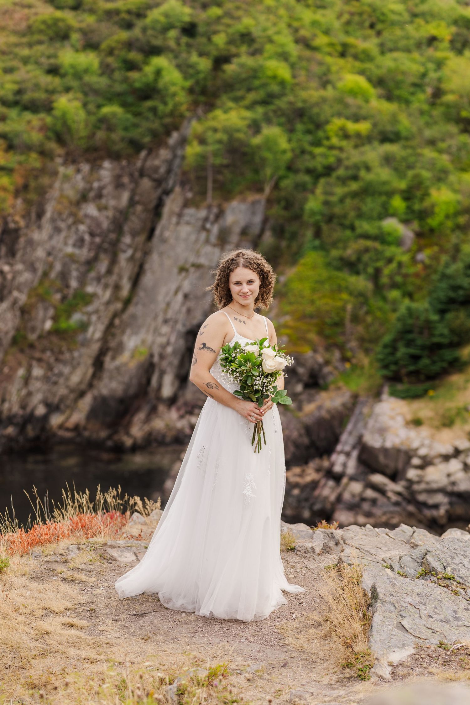 A scenic mountain landscape with a bride in a flowing white dress holding a bouquet against rugged cliffs and greenery.