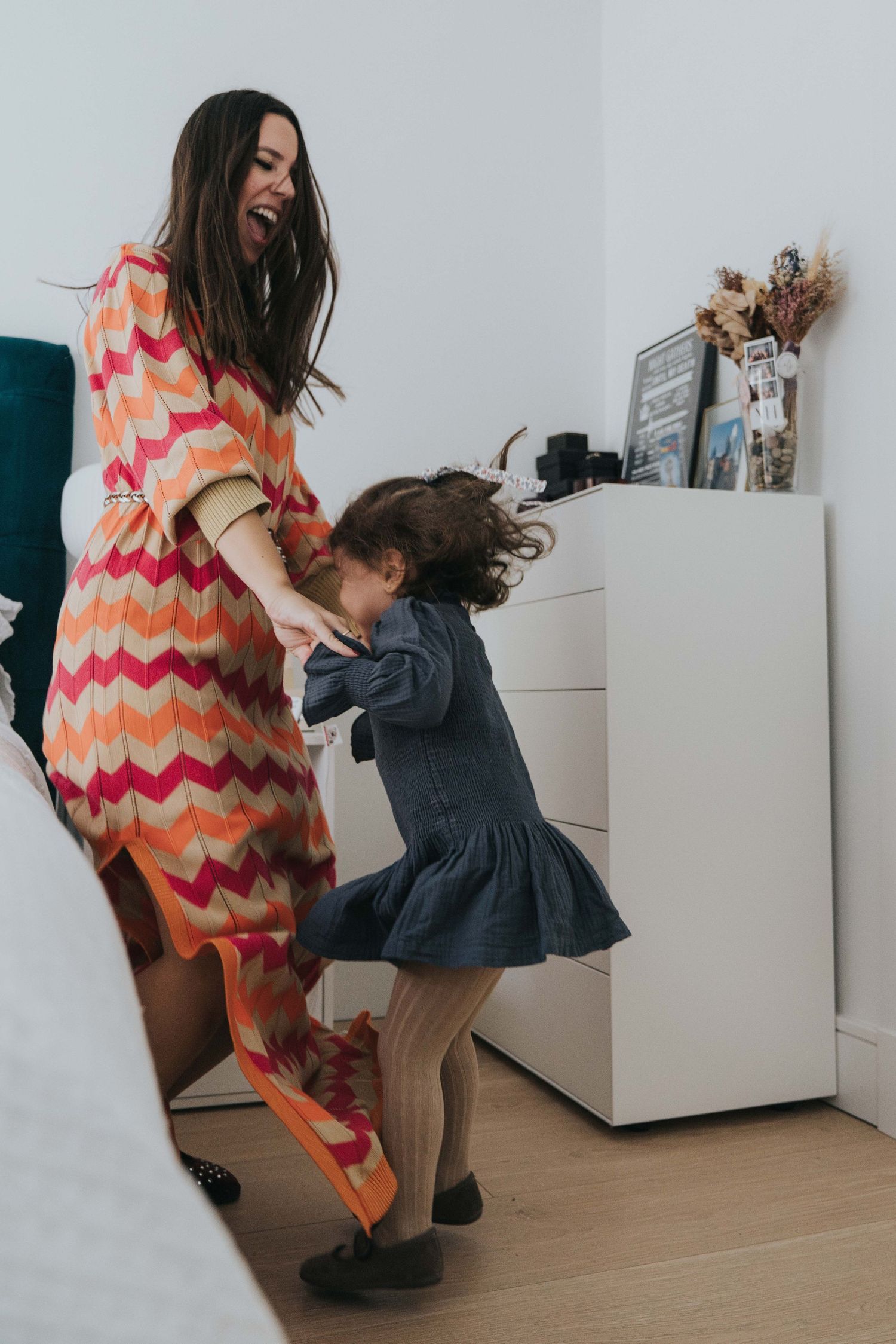 A person in a colorful zigzag dress dances with a child in a denim dress on a wooden floor.