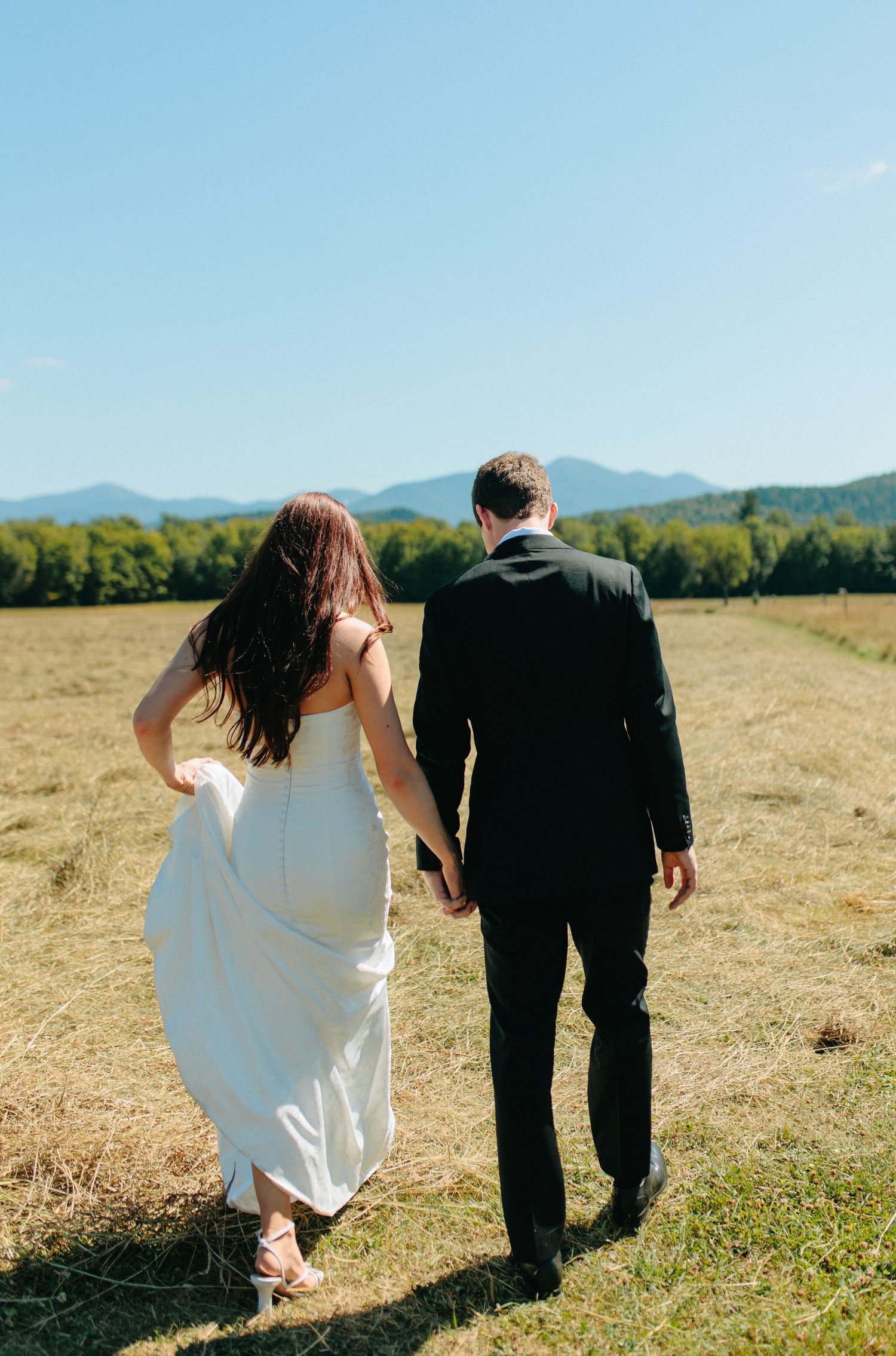 A bride and groom walk together through a golden field with mountains visible in the distance on a sunny day.