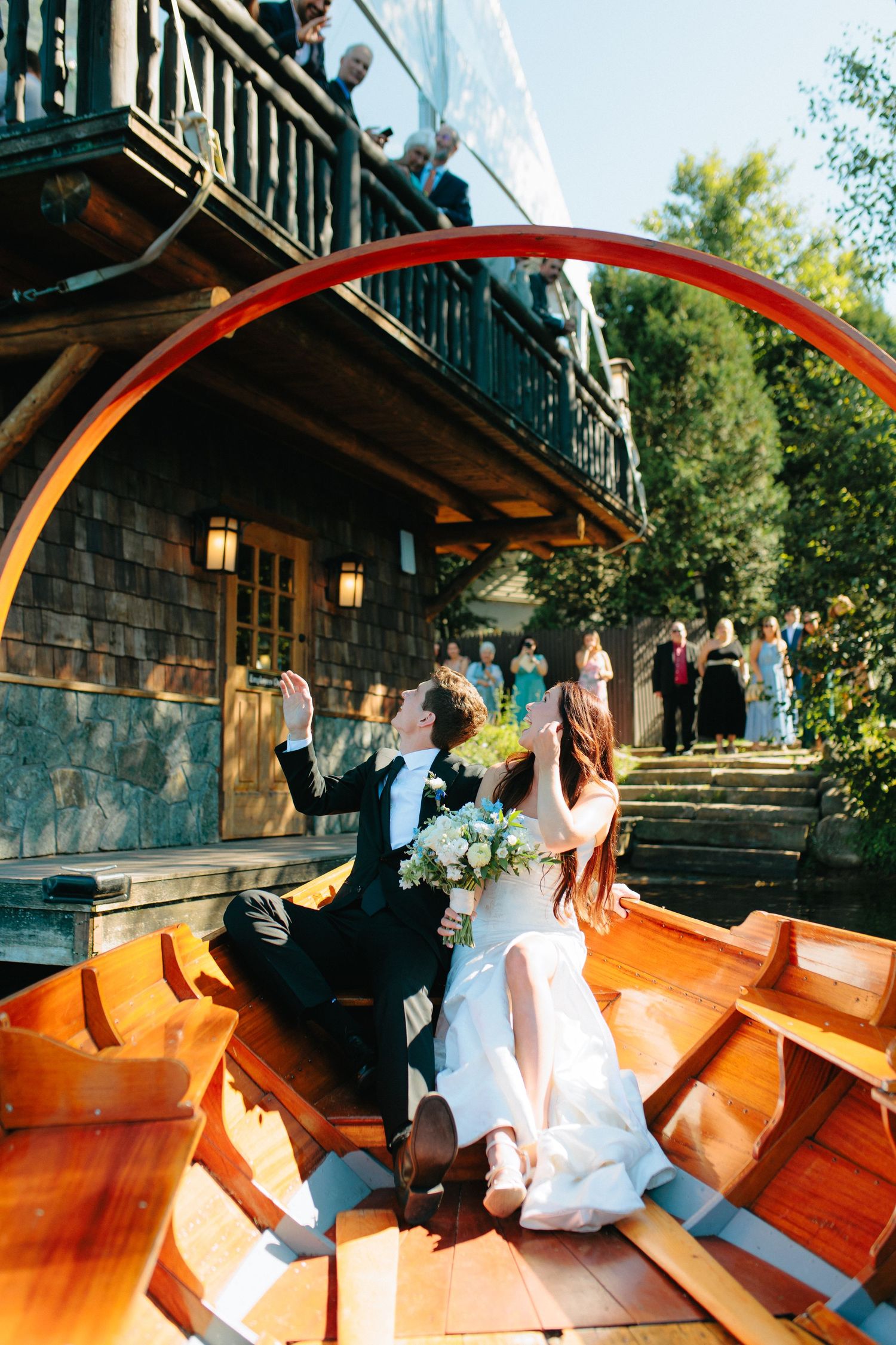 A couple sits in a wooden boat near a rustic lakeside lodge with guests gathered on stone steps and balcony above.