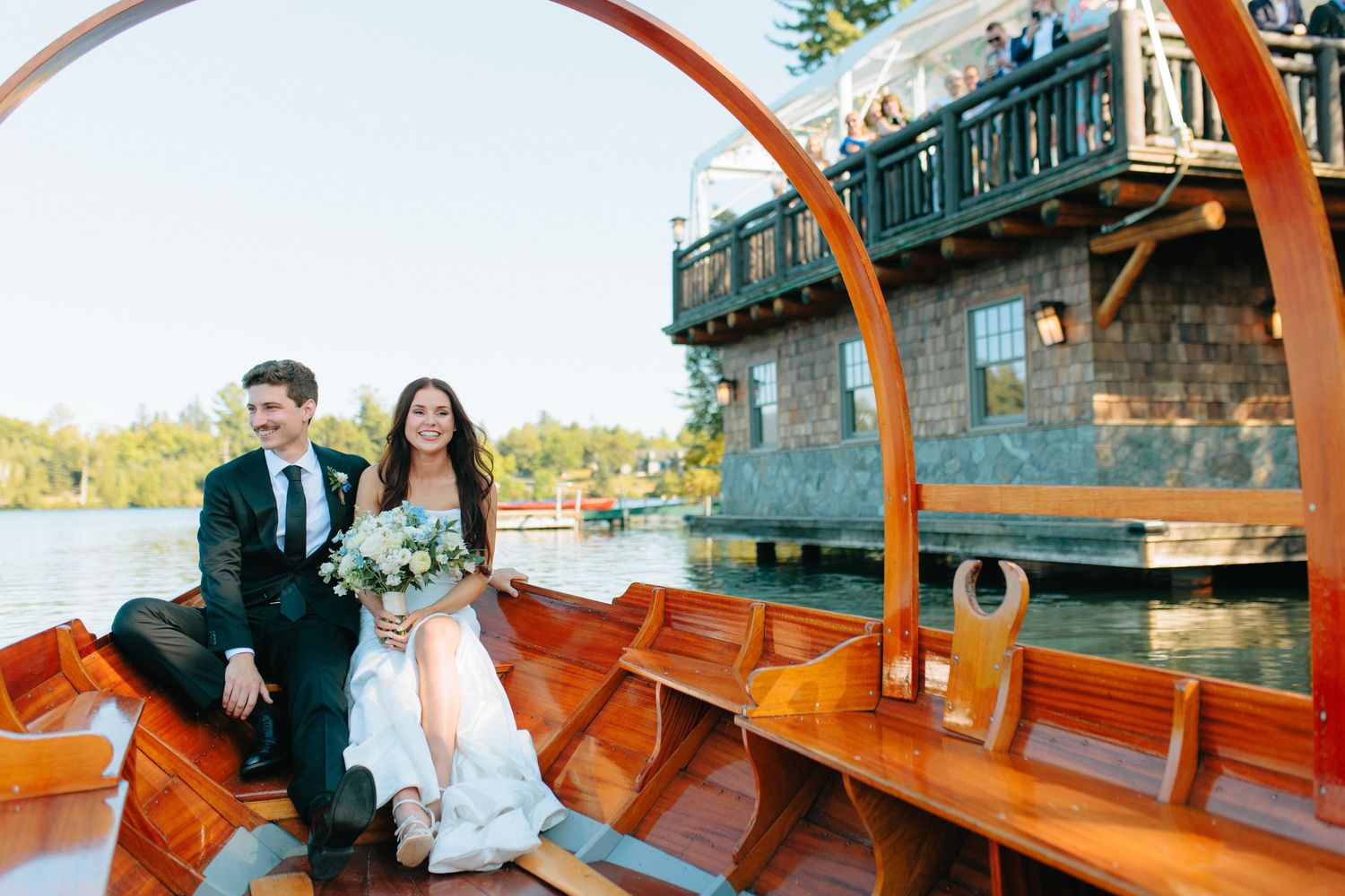A couple sits in a wooden pletna boat on Lake Bled with a traditional waterfront boathouse visible in background.
