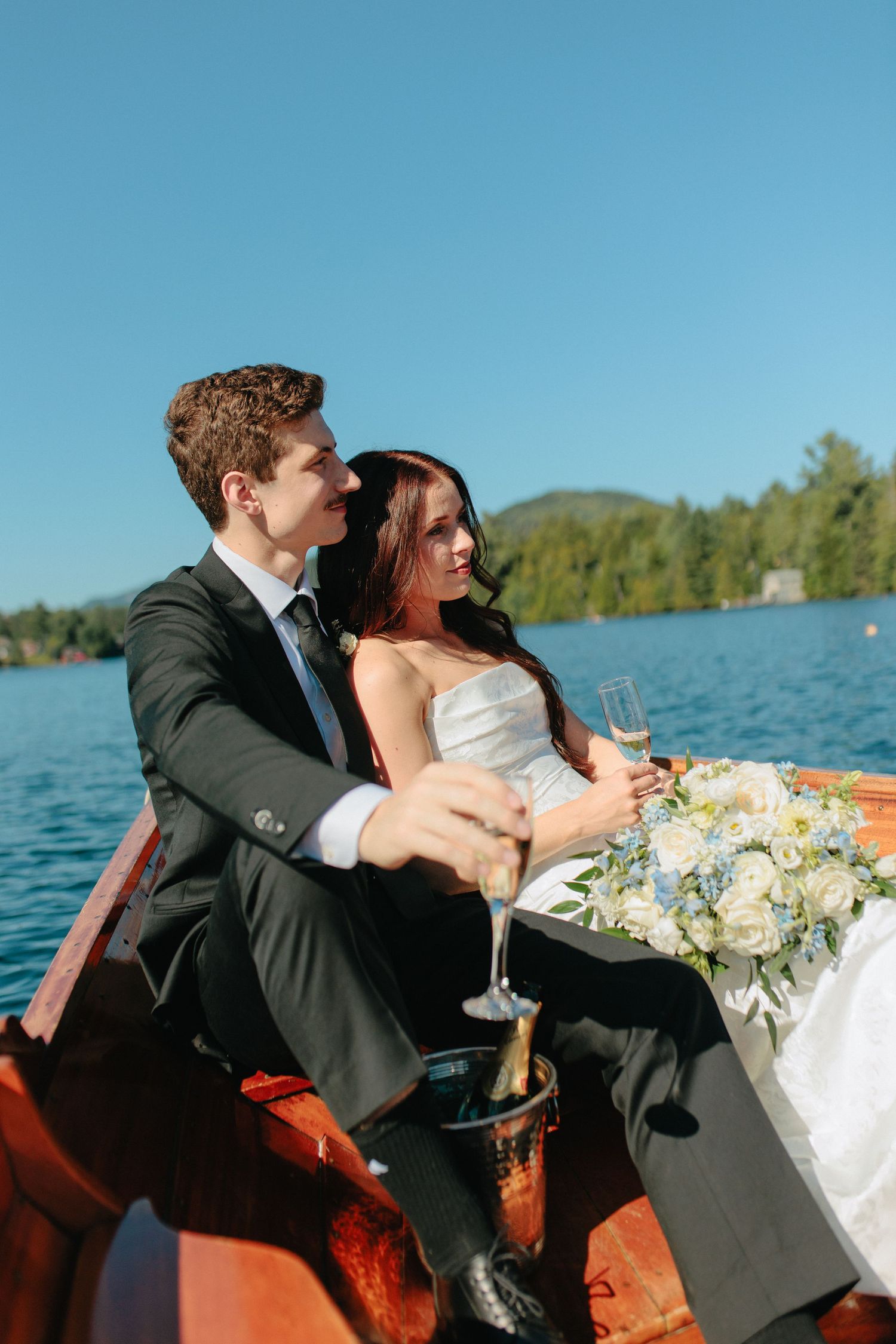 A romantic wedding couple shares champagne while sitting in a wooden boat on a serene lake.