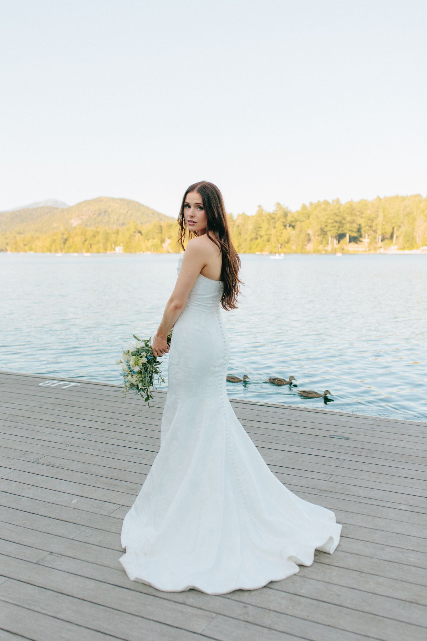 A figure in a white mermaid wedding dress stands on a wooden dock overlooking a serene lake with mountains in the background.