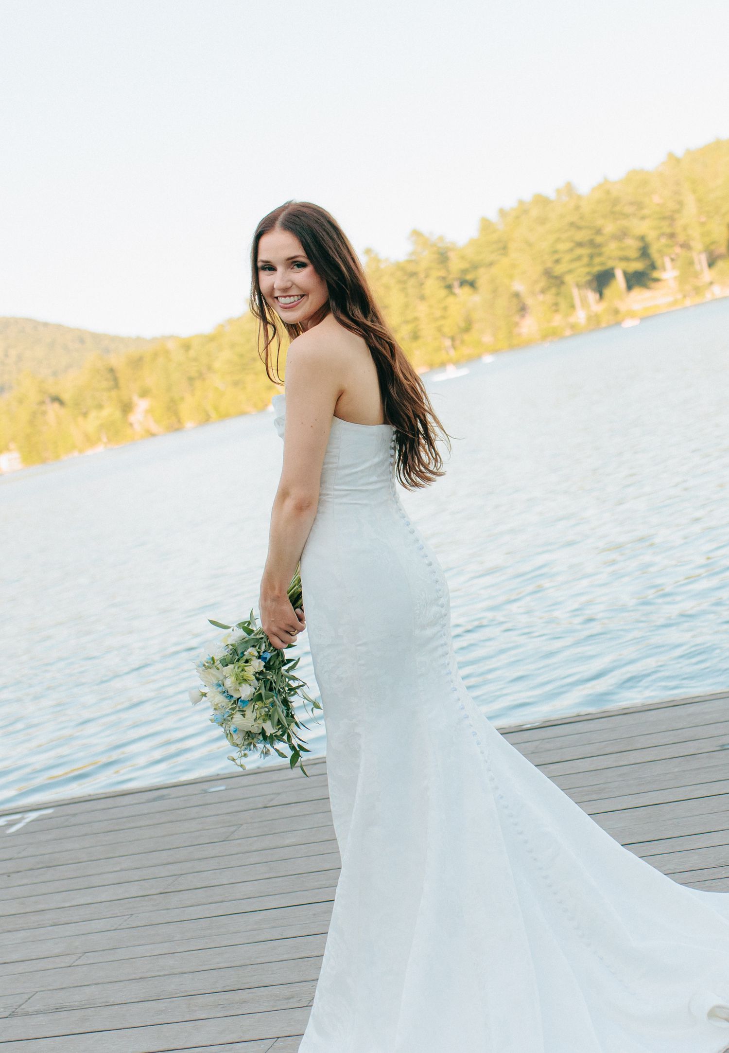 A bride in a flowing white mermaid dress poses on a wooden dock by a lake at sunset.