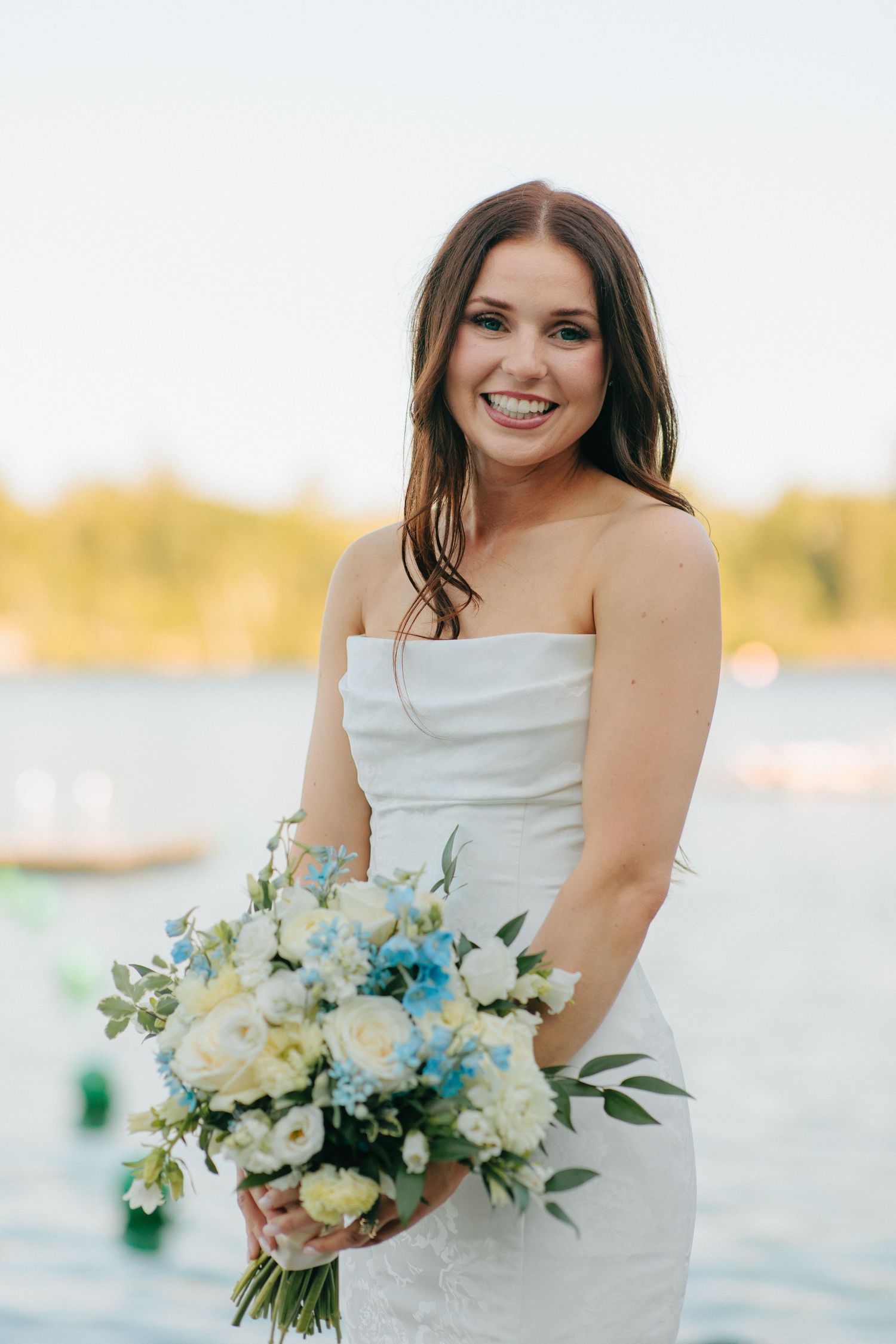 A bride in a white strapless dress holds a beautiful bouquet of blue and white flowers by the water.