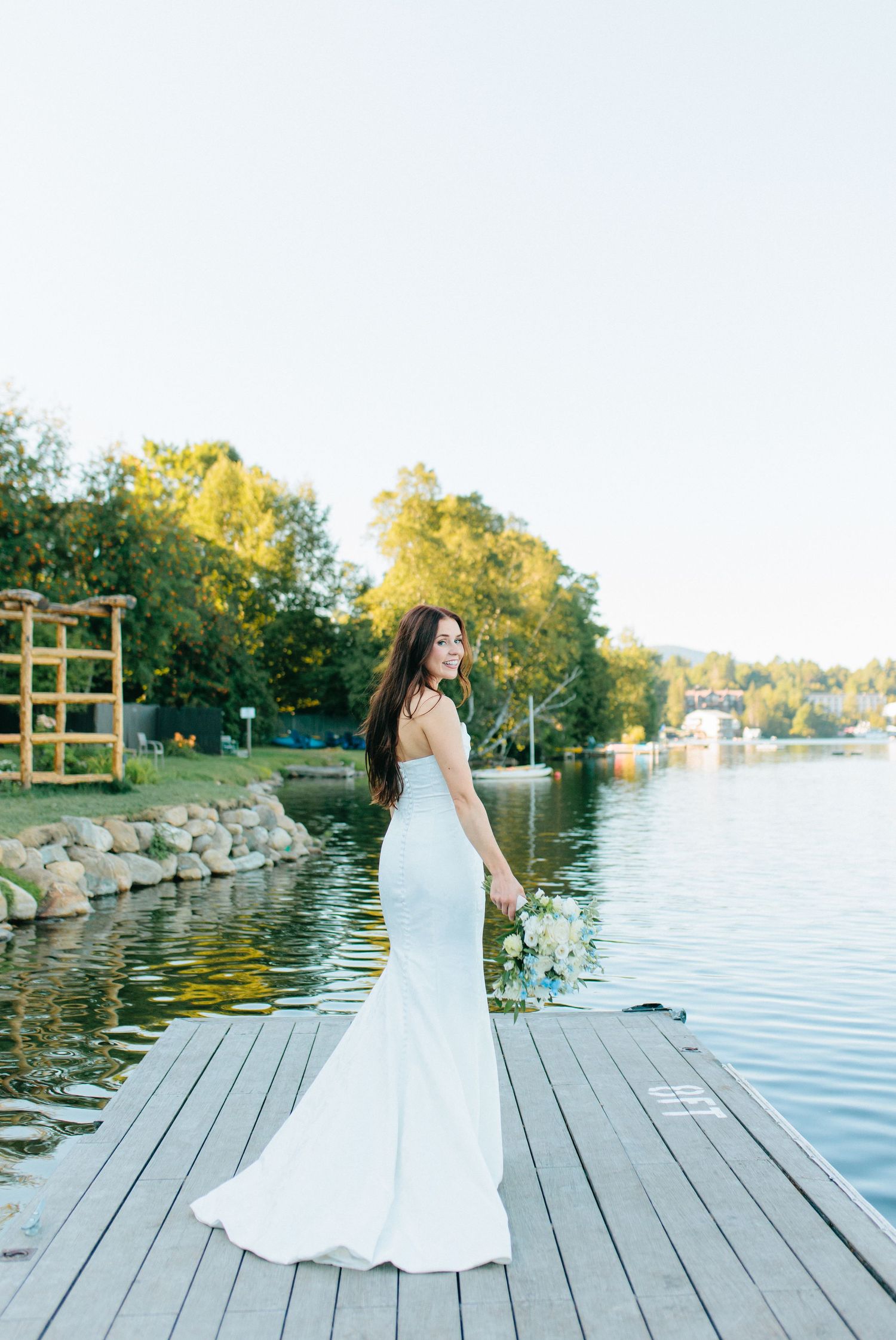 A bride in a sleek white mermaid wedding dress stands on a wooden dock by a serene lake at sunset.