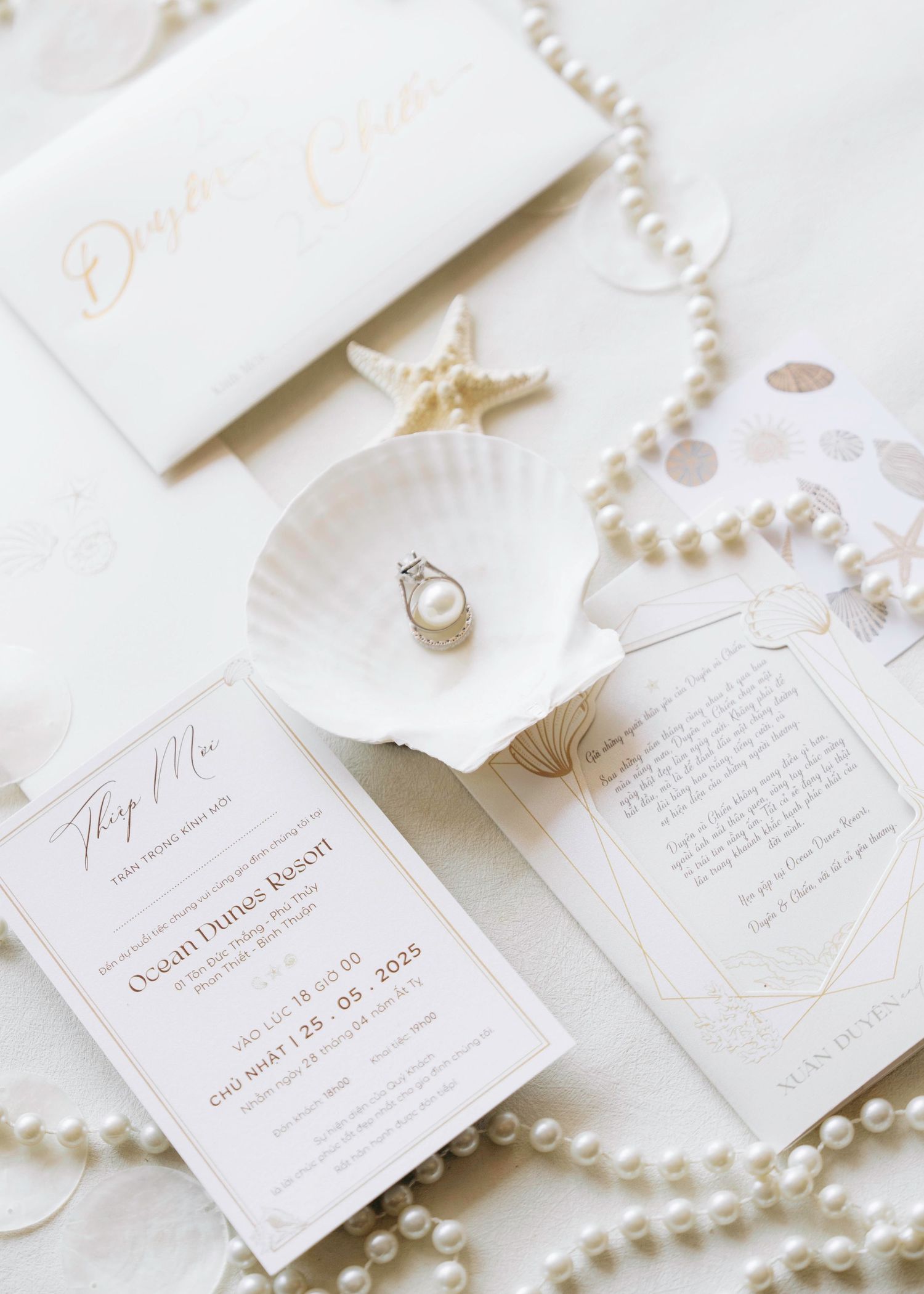 Elegant beach wedding stationery flat lay with pearl ring, seashell, pearl necklace, and ocean-themed invitations.