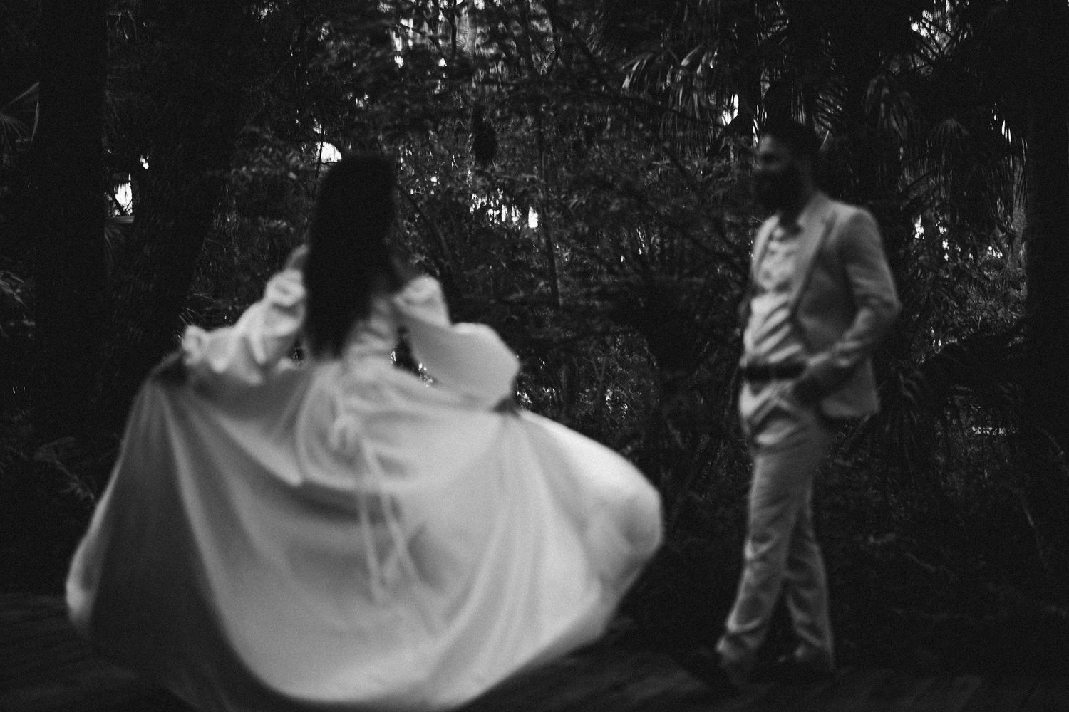 A romantic wedding couple stands beneath string lights in a wooded area during sunset at an outdoor ceremony.