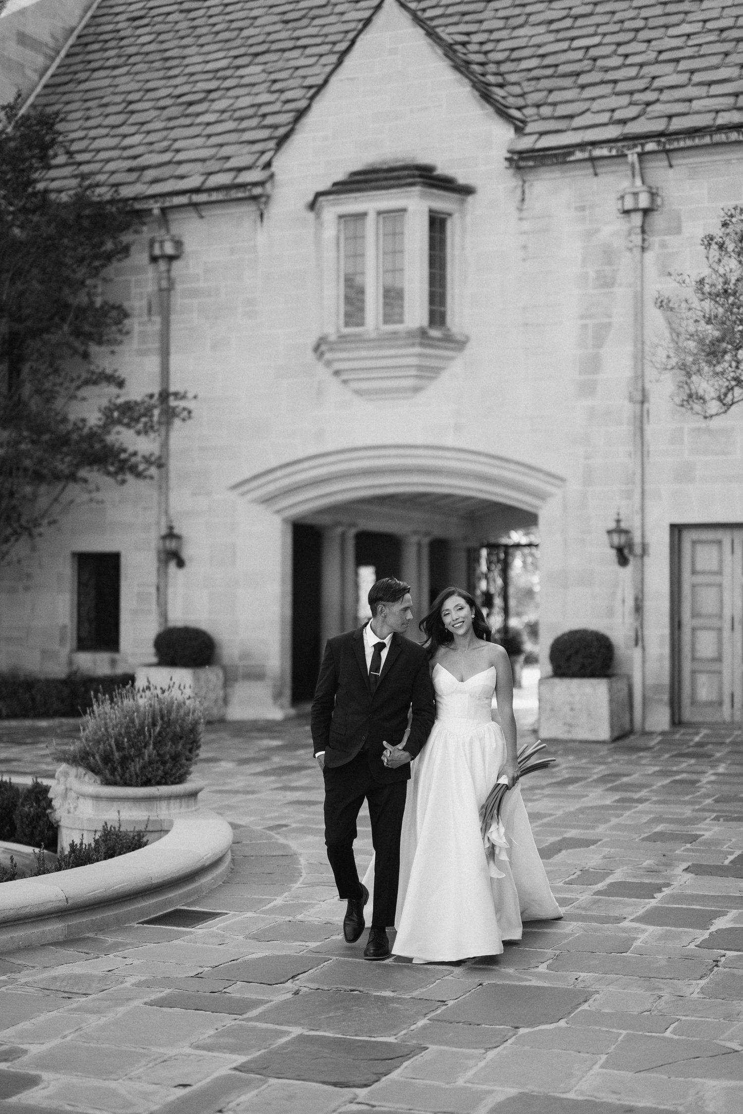 A romantic wedding couple walks along a stone courtyard path beside a historic white building on their special day.