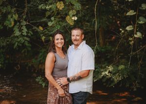 A couple embraces in front of a scenic wooded area with dappled sunlight filtering through the trees.