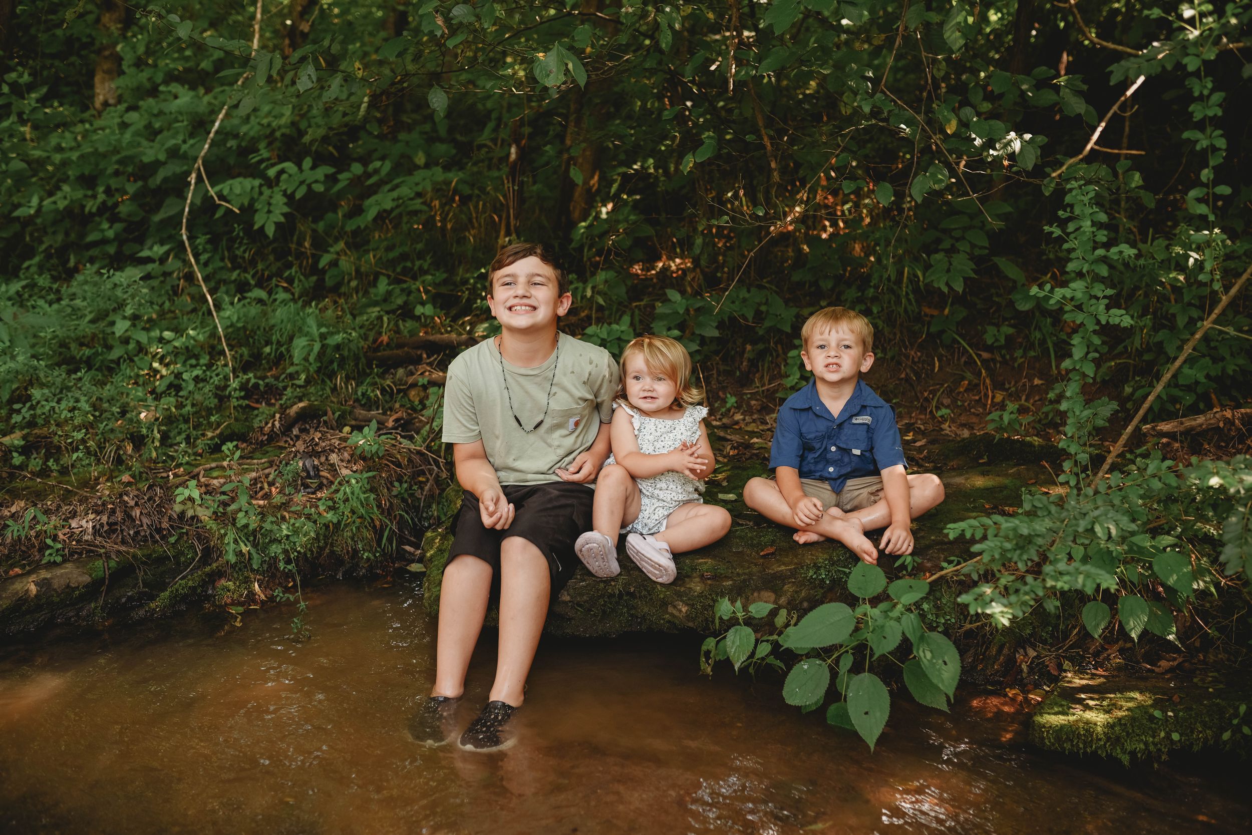 Children sit along a creek bank in a wooded area during summer.