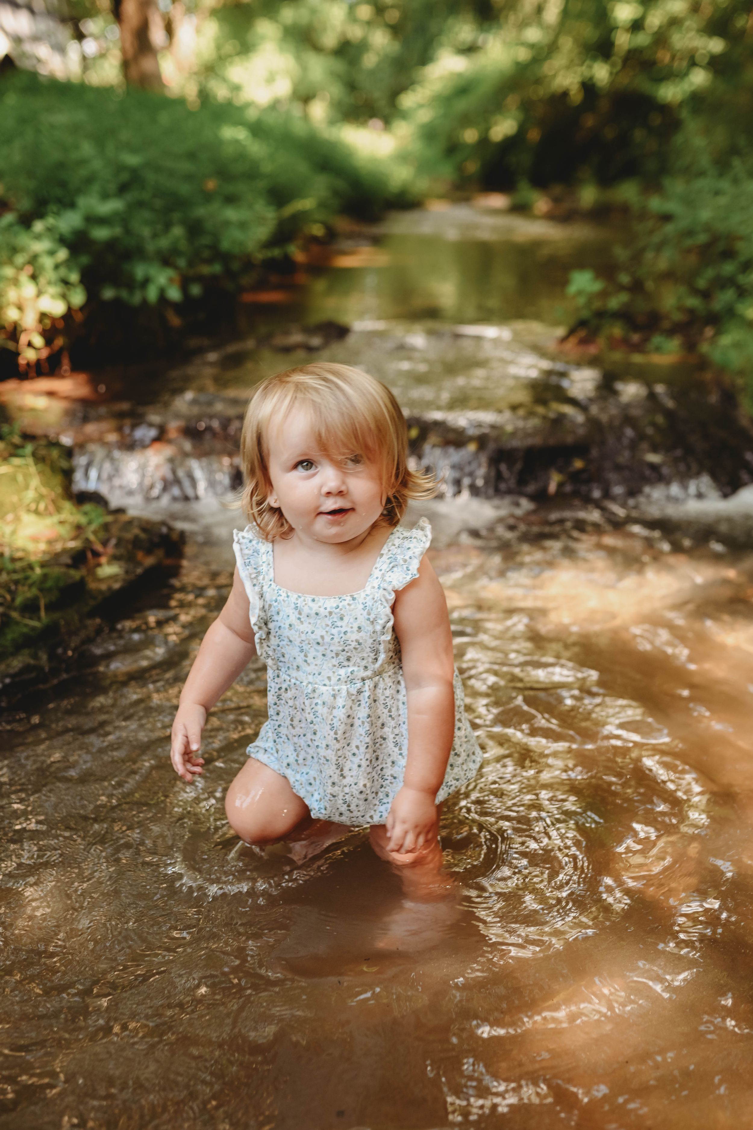 A toddler in a floral dress splashes and plays in shallow creek water.