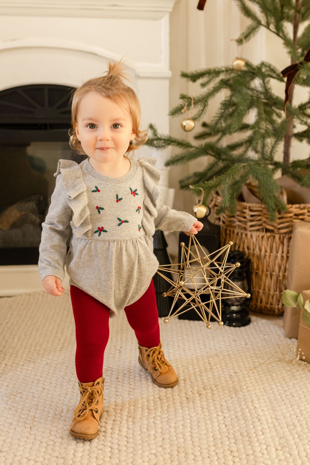 A small child in a gray dress and red tights plays near a Christmas tree on a beige carpet.