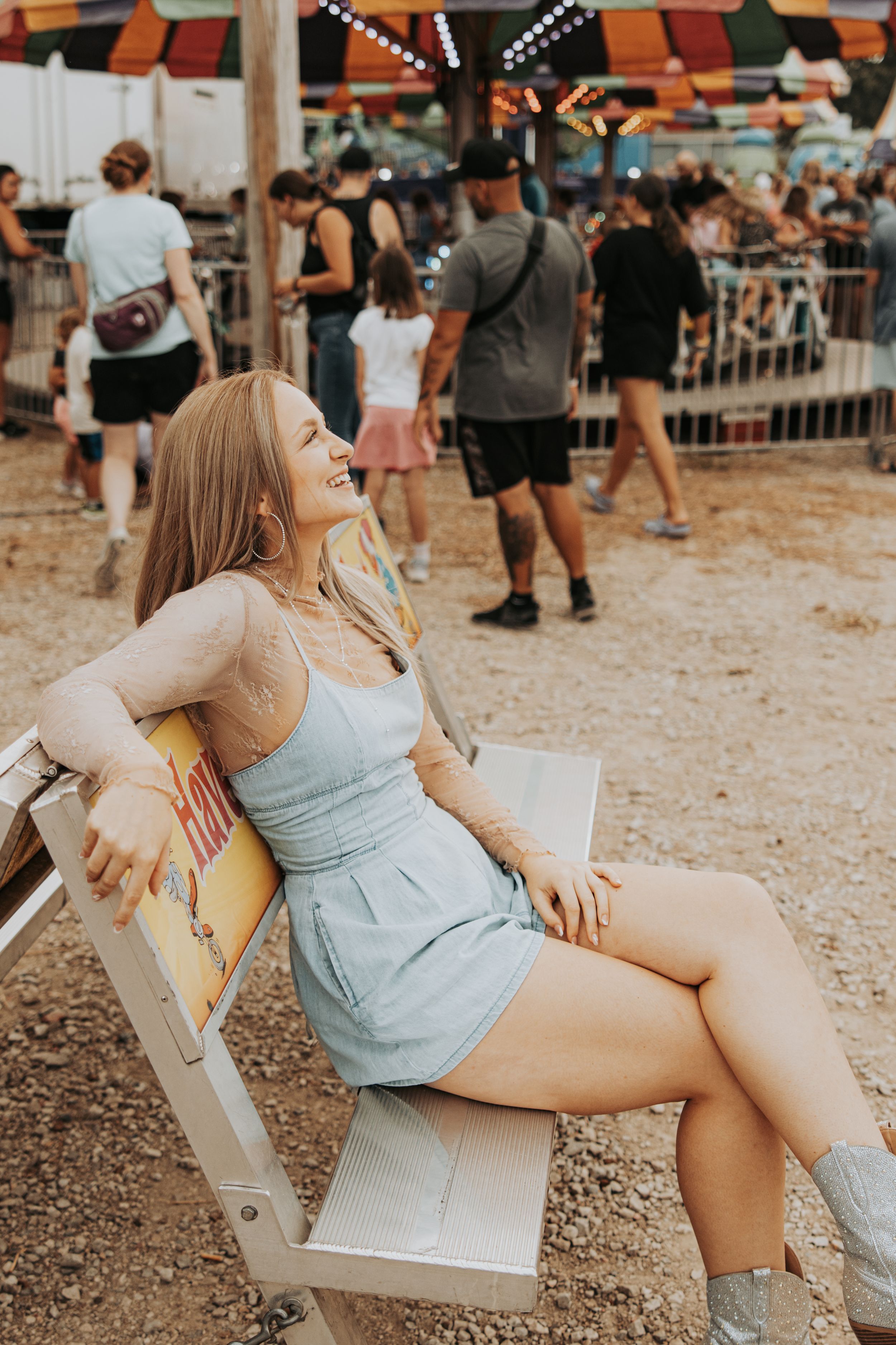 Woman in light blue dress poses playfully on wooden bench at outdoor festival or fair.